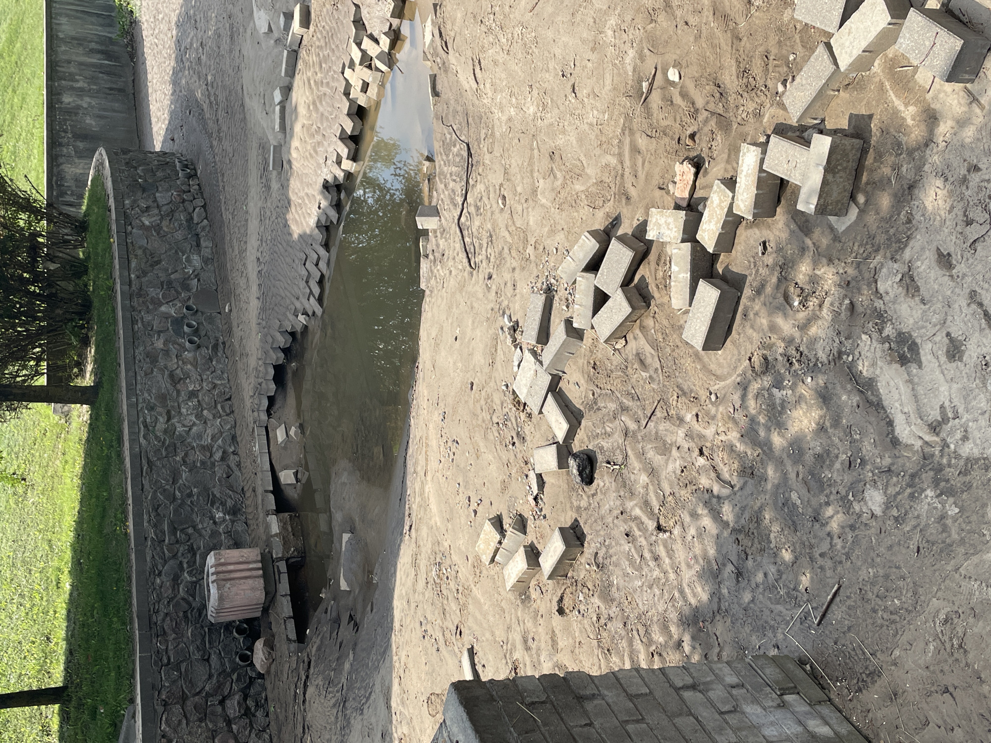 This image depicts a construction site in the city of Hrodna, Belarus, likely a park or public space undergoing renovation. The scene is dominated by exposed earth and sand, with a small pool of water in the center. Scattered across the ground are numerous concrete paving stones, some laid in place and others piled haphazardly. A low stone wall borders a grassy area in the background. There are no people visible in the image. The overall impression is one of disarray and ongoing work.