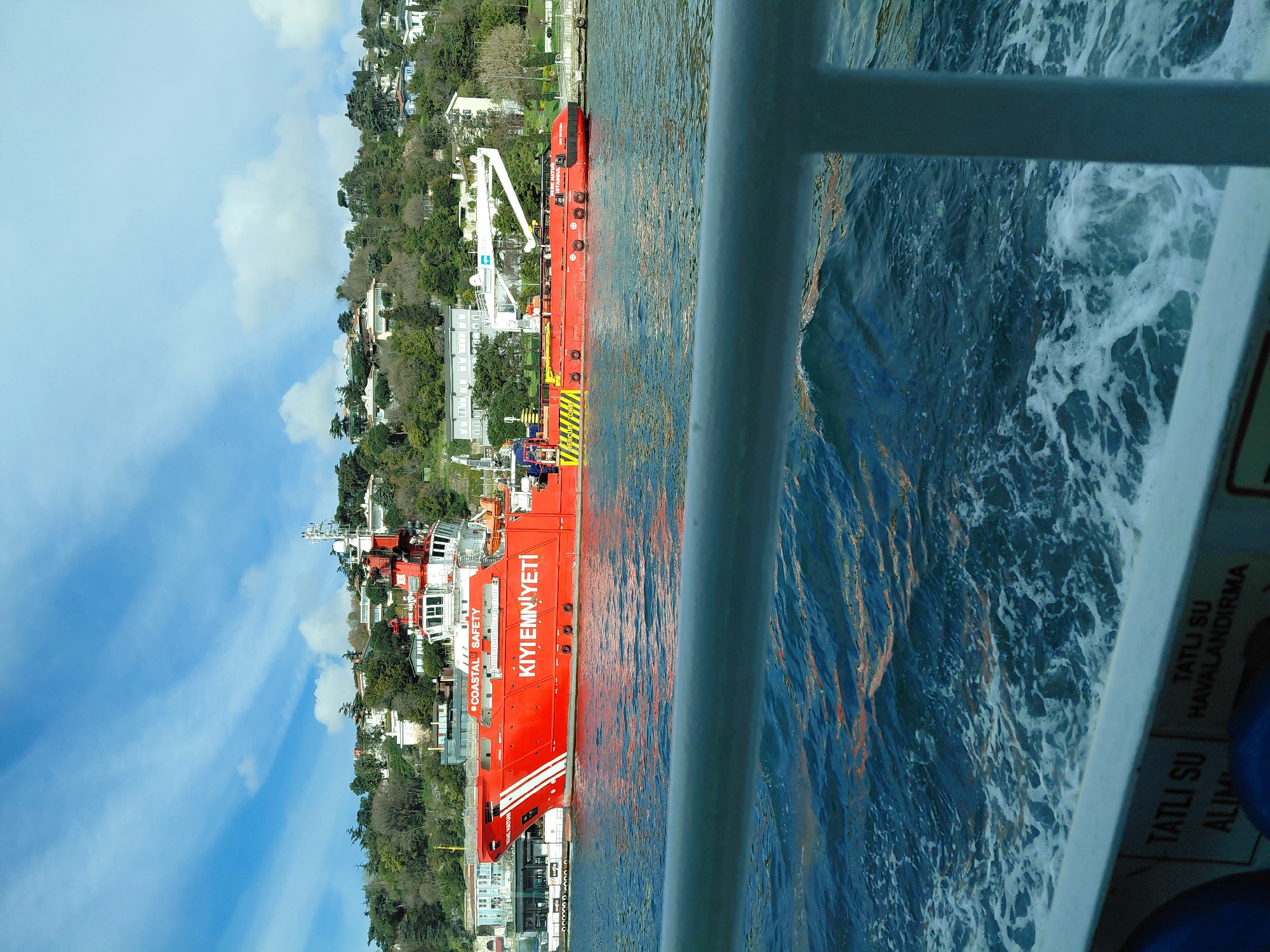 The image shows a view from a moving vessel, with a red and white “COASTAL SAFETY” or “KIYI EMNIYETI” boat docked along the shore. The water is choppy, indicated by visible ripples and whitecaps. The background shows a hillside dotted with buildings and trees. The location is likely Beşiktaş, Turkey, given the text on the boat. A metal railing is in the foreground, partially obscuring the view.