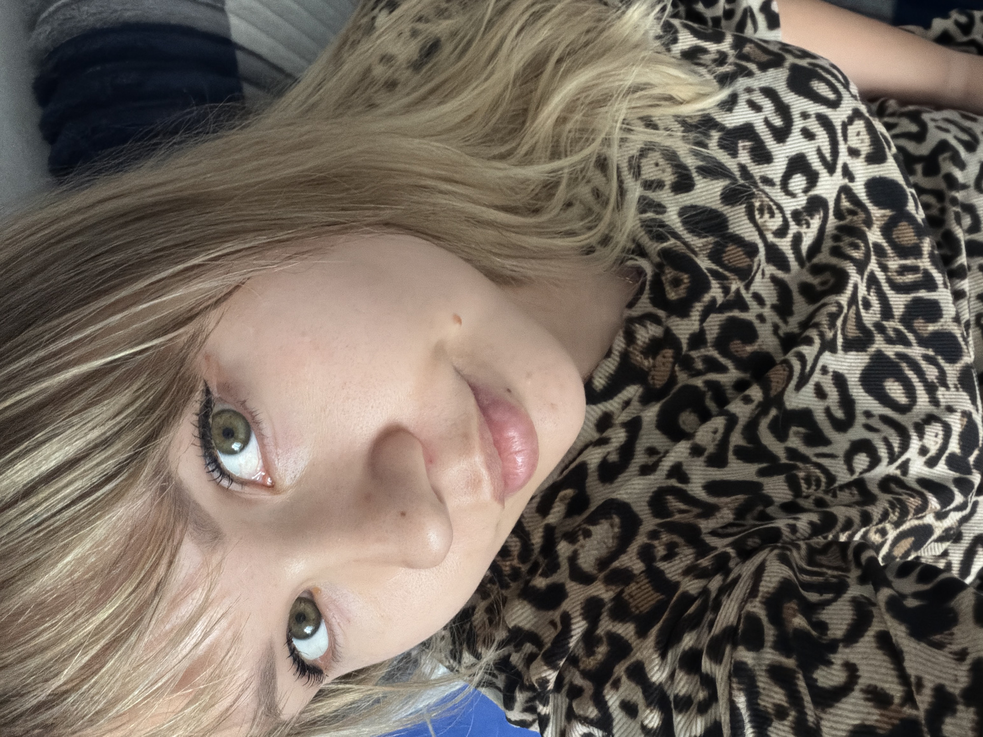 The image is a close-up, angled view of a person with fair skin and light brown/blonde hair. The individual has green eyes and is wearing makeup, including eyeliner and mascara. They are dressed in a garment with a leopard print pattern. The background shows a glimpse of a blue-and-grey striped surface. The location is Unknown, Unknown.