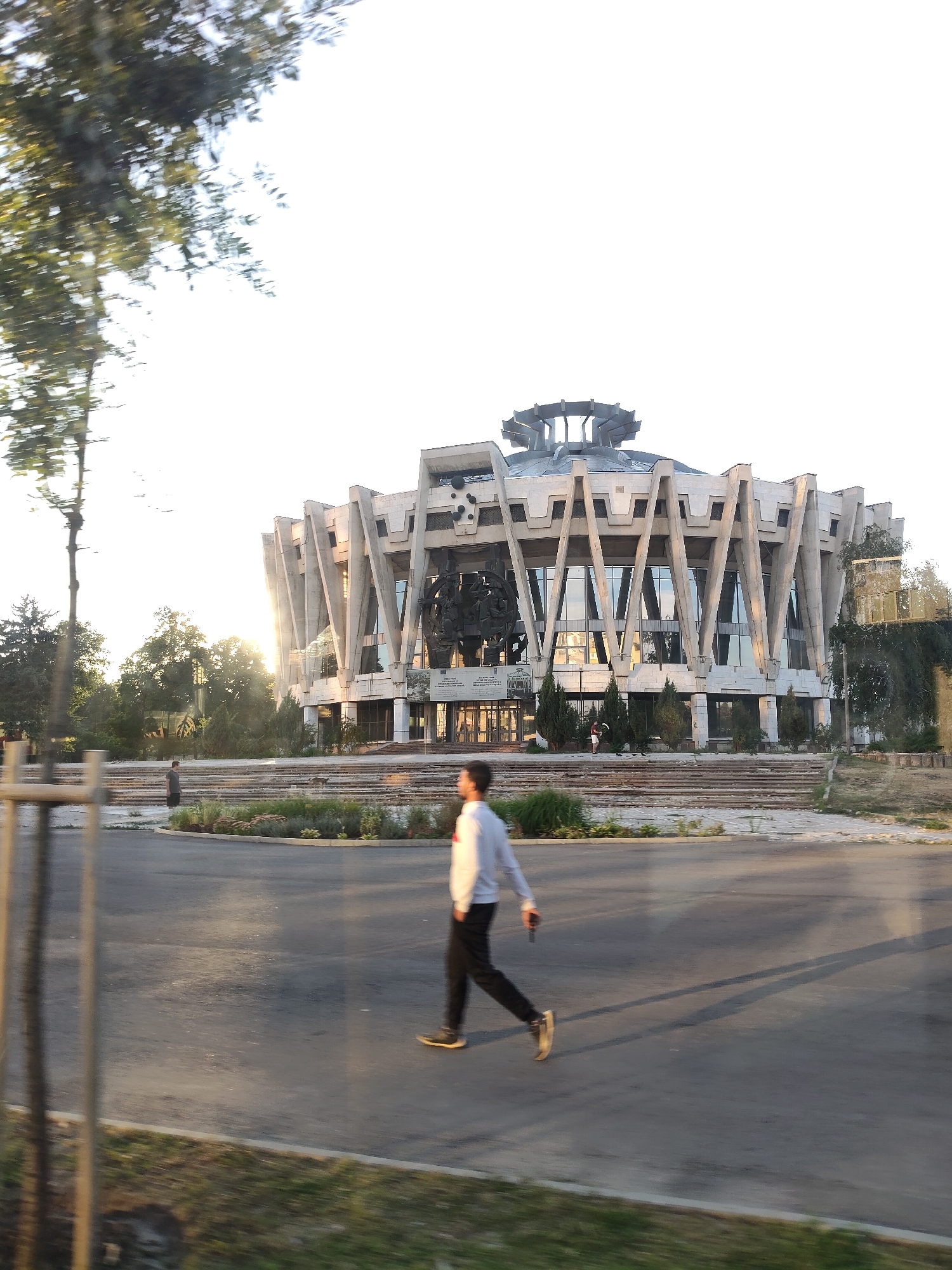 This image depicts the Palace of the Republic in Chișinău, Moldova. The building, a prominent example of Soviet modernist architecture, features a distinctive circular design with a large, stepped roof and a facade characterized by angled concrete elements and extensive glass windows. In the foreground, a young man in a white long-sleeved shirt and black pants is walking from left to right across a paved area, casting a long shadow. The surrounding environment includes trees, greenery, and a road, suggesting an urban setting. The sun is low in the sky, casting a warm light and contributing to the lens flare visible on the building's facade.