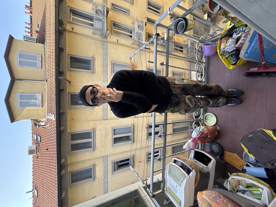 An individual stands centrally on a cluttered residential balcony in a city, consistent with architecture found in Milan, Italy. The person, appearing male, wears a black beanie, black button-up cardigan, camouflage cargo pants, and dark shoes. He is smoking a cigarette, holding it between his right thumb and index finger near his mouth. The balcony floor is tiled in reddish-brown, enclosed by ornate wrought iron railings. Visible on the balcony are multiple lidded plastic containers, some appearing to be for composting or waste, a yellow bag filled with recycling materials, a purple bucket, miscellaneous plastic bags with contents, and a small potted plant. Directly opposite and below the balcony is a multi-story light-yellow building featuring numerous rectangular windows with light gray shutters. This building also displays several small wrought iron balconies and has a terracotta tiled roof with skylights and an exposed air conditioning unit, all beneath a clear blue sky.