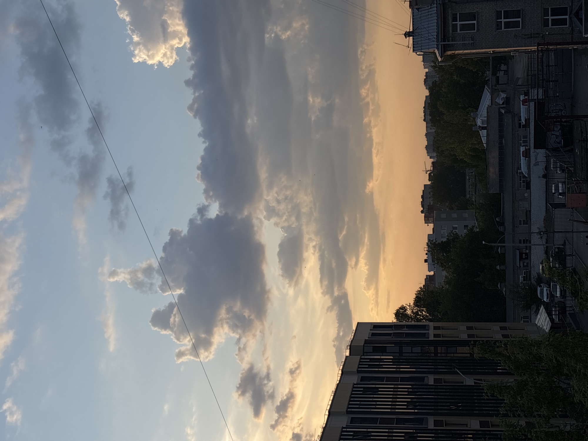 The image captures a cityscape at dusk in Perm, Russia. In the foreground, the side of a modern building with horizontal lines is visible. Above it, a row of buildings and trees creates a somewhat cluttered skyline. In the background, the sky transitions from a light blue to a warm orange color, as clouds are distributed vertically through the frame. An overhead power line cuts across the image diagonally.