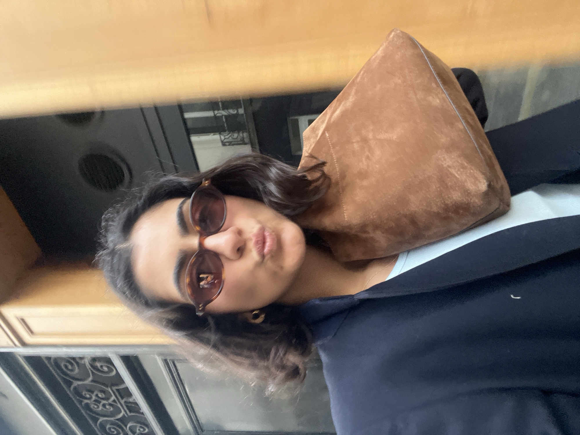 A woman with dark hair and sunglasses poses outdoors. She wears a blue blazer over a light-colored top and has a brown suede bag on her shoulder. In the background is a Parisian balcony with wrought iron detailing and a view of buildings.