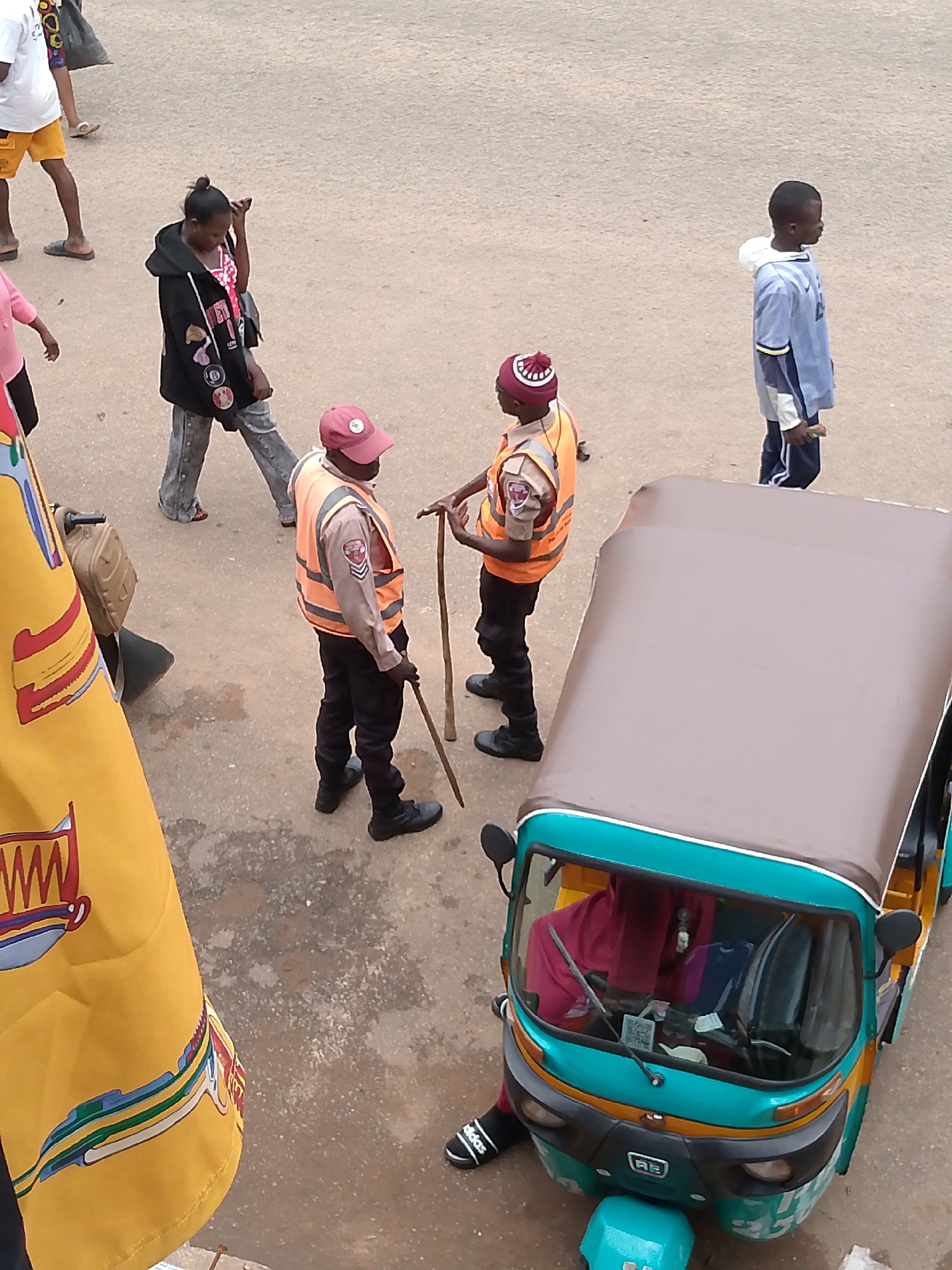 An aerial shot captures a street scene in Bukuru, Nigeria, with several individuals and a three-wheeled vehicle. In the foreground, a bright yellow banner with red, blue, and green designs is partially visible on the left. A teal and yellow auto-rickshaw with a brown canopy is positioned in the lower right quadrant, with a person wearing a red headscarf driving it. Two individuals in orange vests and dark pants stand near each other; one holds a wooden stick. Another person, a young boy in a blue and white jacket and dark pants, stands to the right, looking away from the camera. Further to the left, a woman in a black hooded jacket and jeans walks away from the viewer, while another individual in a white t-shirt and yellow shorts is seen from the waist down on the far left. The ground appears to be a paved surface with some patches of dirt and debris.