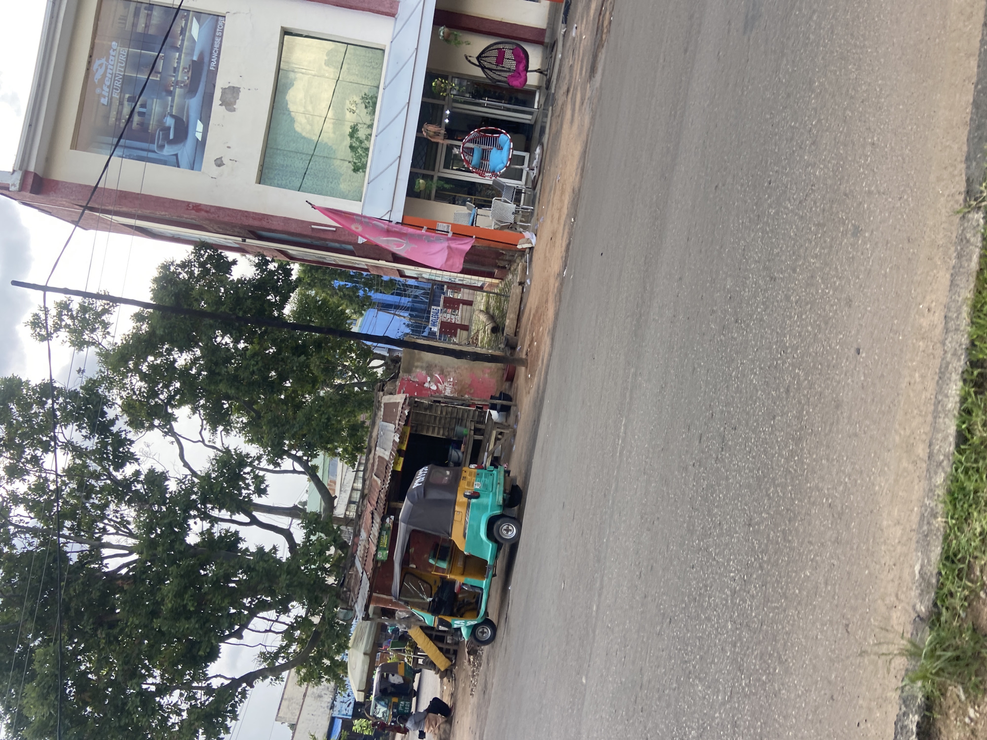 The image depicts a street scene in Jos, Nigeria. In the foreground, a paved road runs diagonally from the lower left to the upper right corner. On the right side of the road, a multi-story building with a white facade and red trim is visible. This building features large windows, some of which reflect the sky. A banner with the text "Lifemate FURNITURE" and an advertisement for furniture are displayed on the upper part of the building. Below this, a franchise store is advertised. A pink flag is planted in front of the store, and outdoor seating with chairs and tables is set up. Two hanging chairs, one blue and one pink, are also displayed outside.
To the left of the building, a small shack with a corrugated metal roof stands beside the road. A turquoise and yellow auto-rickshaw, commonly known as a "Keke Napep," is parked in front of this shack. The auto-rickshaw has a black canopy. Further to the left, another auto-rickshaw is partially visible. A person wearing a white shirt and brown pants is seated on the ground to the left of the shack. Behind the shack and to the left of the building, large trees with dense green foliage dominate the scene. Overhead power lines are visible crisscrossing through the branches of the trees and above the street. The overall environment appears to be an urban or semi-urban area with commercial activity and roadside vendors. The sky is partly cloudy.