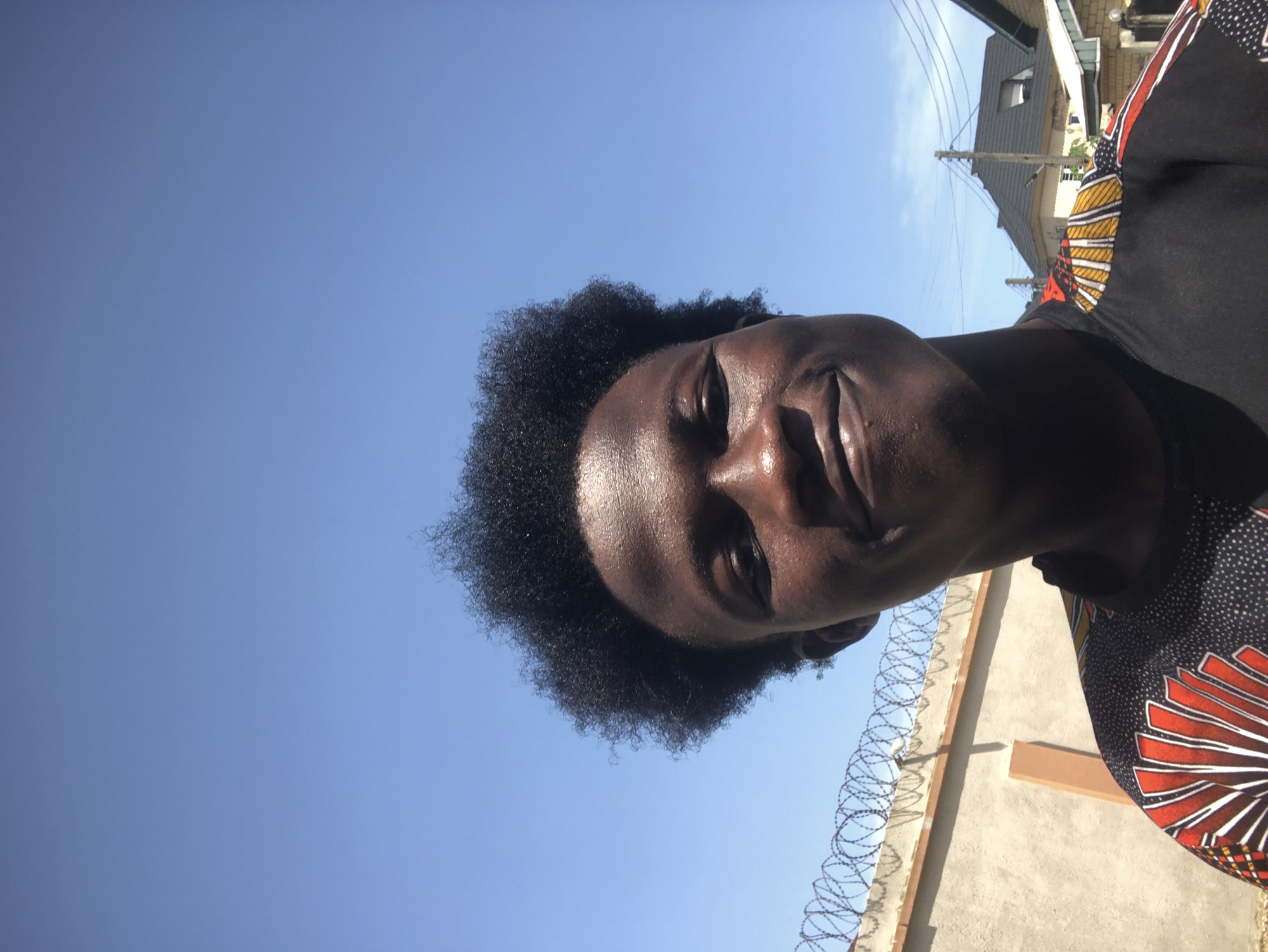 The image captures a low-angle portrait of a person with dark skin and curly black hair, looking upwards and smiling warmly. The strong, direct sunlight highlights their forehead, cheekbones, and nose, indicating a bright, clear day. The person is wearing a black top adorned with a vibrant, radial pattern in red, yellow, and white, suggesting a traditional African print.
The setting is outdoors in what appears to be a residential or urban area in Ado, Nigeria. The background is dominated by a clear, bright blue sky. To the upper right, elements of buildings with dark roofs, utility poles, and overhead power lines are visible against the skyline. To the lower right, a light-colored concrete wall topped with coiled razor wire runs across the frame, with distinct shadows cast upon it by the bright sun. The overall impression conveys a cheerful moment captured in a sunny, outdoor environment. There is no visible text in the image.
