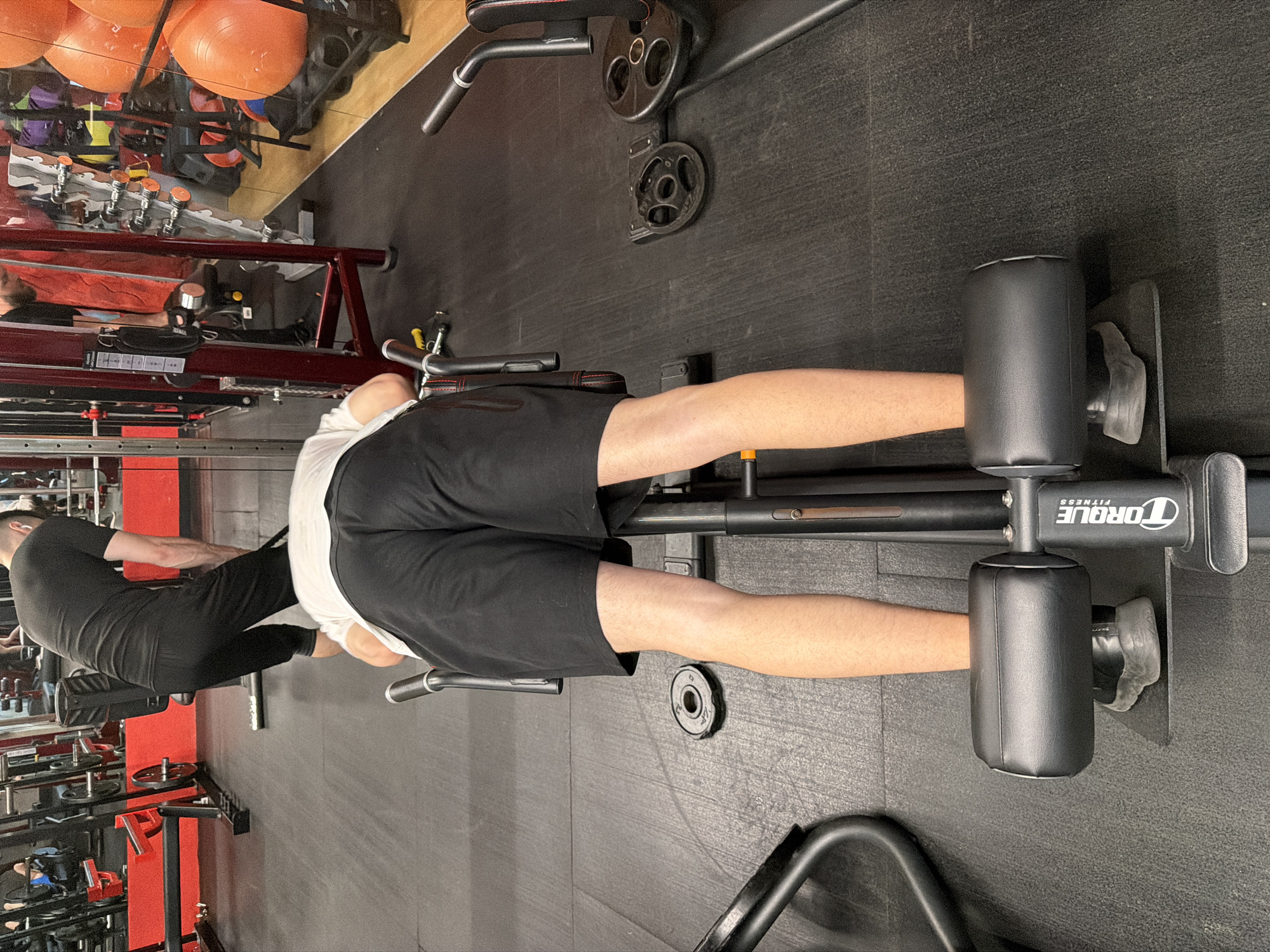 A young man with dark hair, wearing a black t-shirt and black shorts, is performing a hyperextension exercise on a black and red workout machine in a gym. His body is positioned horizontally, with his legs secured by padded restraints. His face is not visible as he is facing down. In the background, another person in a black t-shirt and black pants is seen assisting with a weight plate, and various gym equipment, including weight racks, dumbbells, and exercise balls, are visible. The gym floor is dark gray and appears to be made of rubber matting.