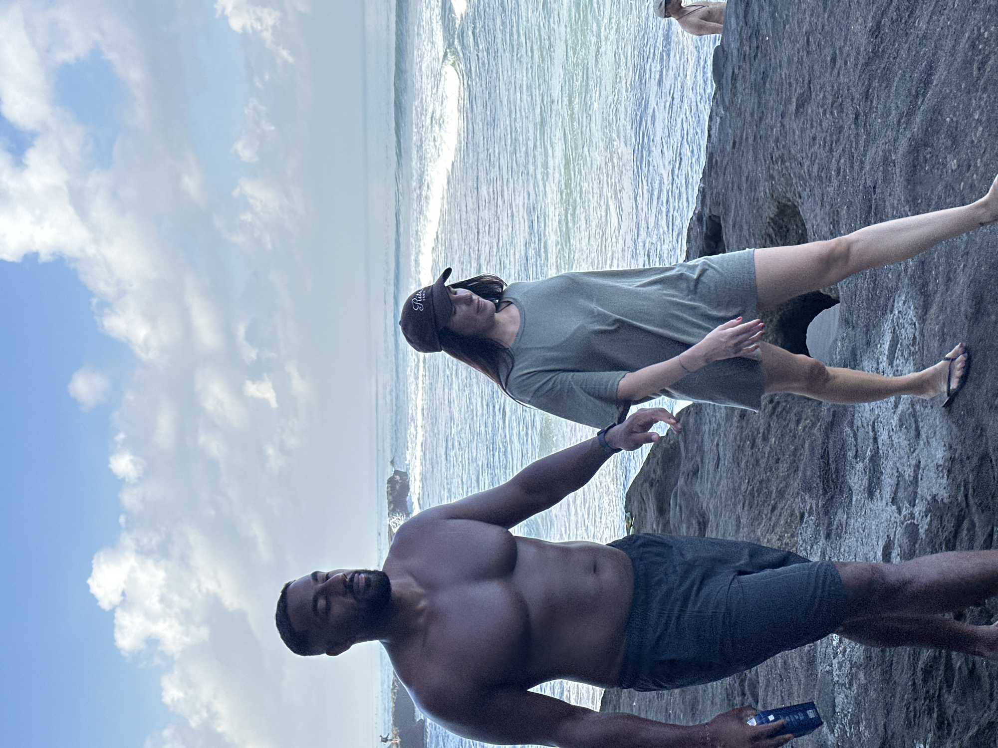 Two individuals walk along a rocky coastline in North Kuta, Indonesia, with the ocean and sky in the background. The person on the left, a muscular man with dark skin and a beard, is shirtless and wearing dark shorts. He is holding a blue object in his left hand. The person on the right, a woman with long dark hair, is wearing a loose olive-green t-shirt and a baseball cap. She is looking down as they walk. The environment is characterized by dark, textured rocks leading to the water, with gentle waves rolling in. The sky is a mixture of blue and white clouds. In the distance, another person is visible standing in the water.