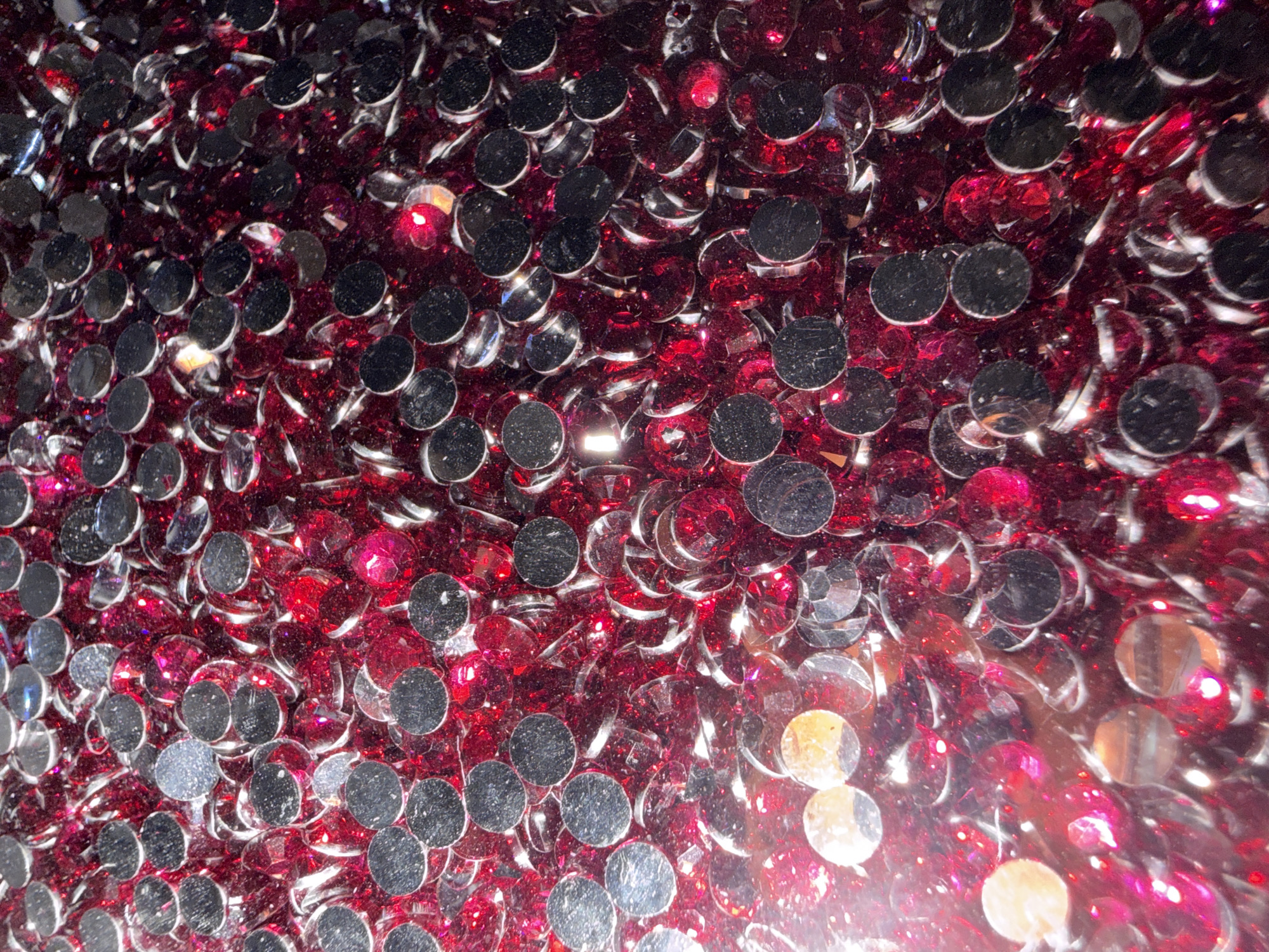 The image is a close-up, overhead shot of a pile of small, flat, round, silver-backed sequins mixed with faceted red gems. The sequins have a reflective, metallic surface, while the gems are translucent and faceted, creating a sparkling effect. The lighting is bright, with some areas appearing overexposed and blown out, particularly on the left side of the image, creating a hazy glow. This suggests the gems and sequins are likely used for decorative purposes, such as crafting, embellishments, or in costumes. There are no people, animals, or recognizable landmarks visible. The image is devoid of any context that would place it within a specific city or location like Portland, United States of America.