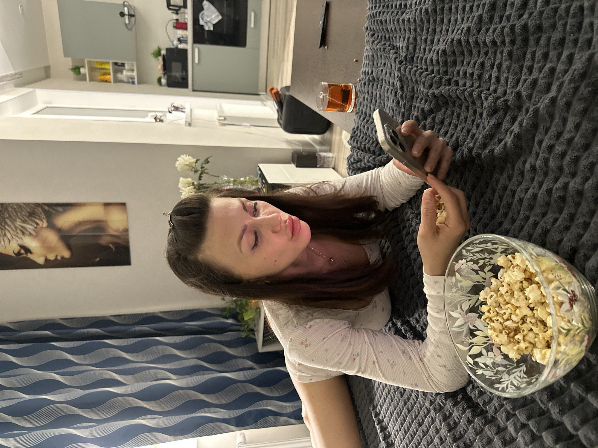 A woman is lying on a textured gray bedspread in what appears to be an apartment. She is wearing a white long-sleeved floral shirt and looking at her smartphone. In her other hand, she holds a piece of popcorn. A large glass bowl of popcorn sits next to her on the bed. In the background, there is a blurred view of a modern kitchen with light gray cabinets. A glass of amber liquid sits on a surface near the head of the bed. The blue-and-white patterned curtain suggests this is not outside. Based on the presence of a modern apartment interior and common items, the photo likely depicts a relaxed, domestic scene, however, the image lacks geographic detail so cannot confirm the exact location.