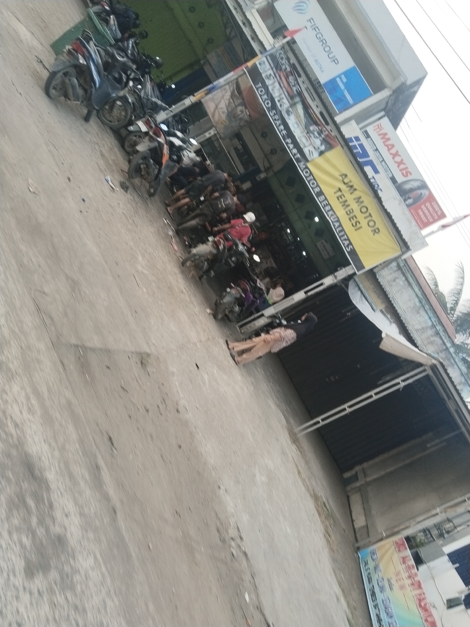 The image shows a street scene with several parked motorcycles in front of a store. The store has signage, including "FIFGROUP" and "MAXSIS", suggesting it might be a motorcycle dealership or service center. There are also banners with text in Indonesian, one of which reads "AJM MOTOR". A few people are visible, some standing and others appearing to be working or browsing at the store. The environment appears to be an outdoor urban setting. The ground is paved, and there are buildings in the background. The overall impression is that of a busy commercial area.
