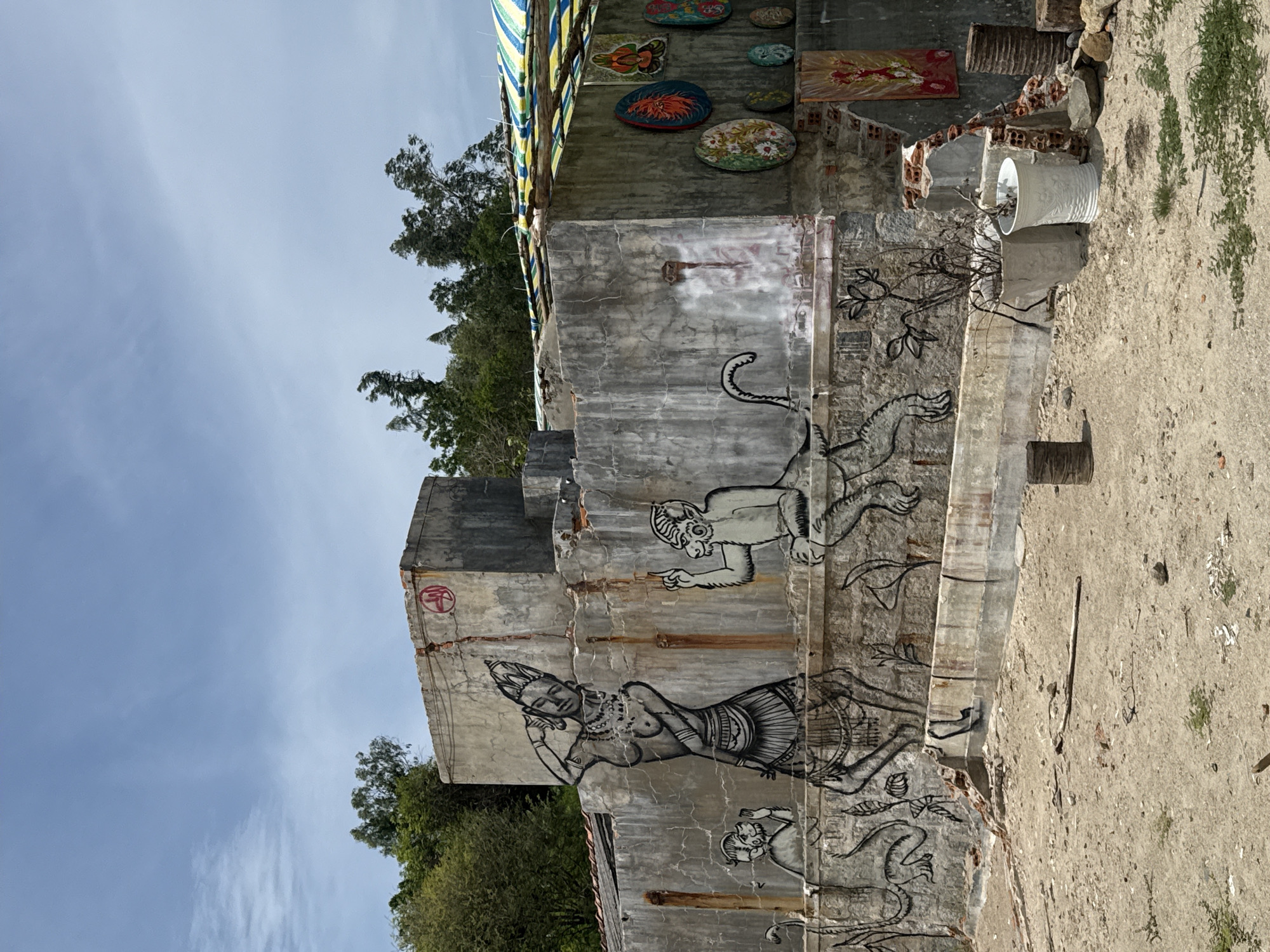 The image depicts an outdoor scene in Vĩnh Lương, Vietnam, featuring a distressed concrete wall adorned with black and white murals. The murals showcase figures of a woman with ornate headdress and jewelry, and several monkeys in various poses. One monkey appears to be reaching up towards the woman, while another is seated and gesturing with a finger. To the right of the main mural, a smaller, seated monkey is depicted. The wall itself is weathered, with visible cracks and patches of rust staining.
Above the murals, on a higher section of the wall, a red circular symbol with a coiled design is present. To the right of the main wall, a section of the structure is covered by a striped tarp, made of blue, green, and yellow fabric, supported by wooden poles. Attached to this section of the wall are several round, colorful art pieces with floral motifs, as well as a rectangular painting featuring a red and gold figure.
The ground in front of the wall is sandy and uneven, with scattered debris, small rocks, and sparse green vegetation. In the foreground, there are several potted plants, one appearing dry and barren, placed in concrete pots. To the far right, wooden cylindrical objects and larger rocks are visible. The background shows a dense cluster of green trees under a bright, partly cloudy sky. The overall impression is one of an artistic, somewhat dilapidated outdoor space.