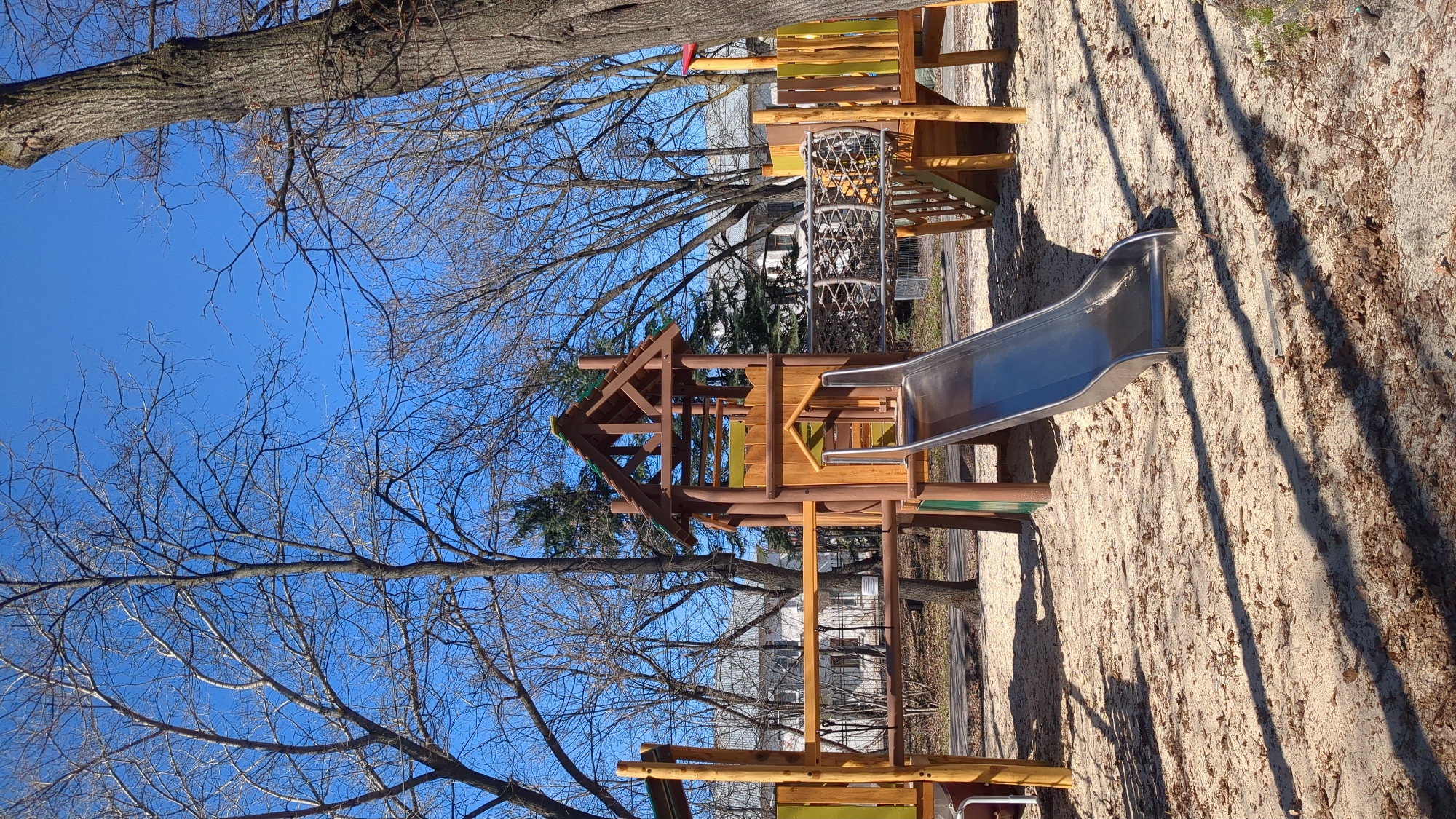 The image shows a children's playground in Ulyanovsk, Russia. The playground equipment is made of wood and metal, including a slide, a climbing structure with a net, and a multi-level playhouse-like structure. The ground is covered in sand and scattered leaves. Bare trees surround the playground, with a clear blue sky visible above. In the background, residential buildings can be seen. No people are visible in the image.