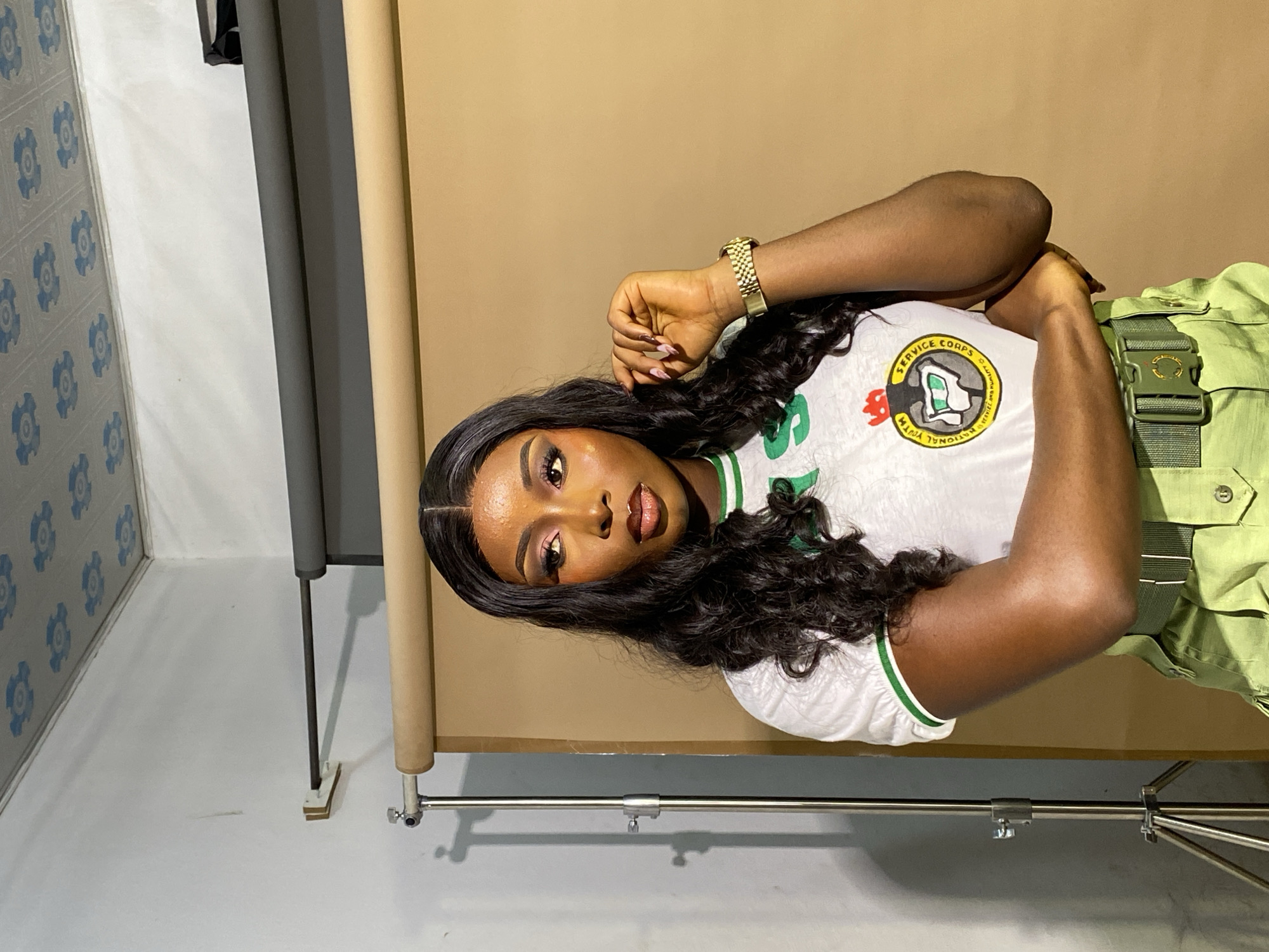 A young Black woman with long, dark, wavy hair and full makeup poses in a studio. She is wearing a white t-shirt with green trim and a circular emblem on the left chest, and olive green pants with a matching belt. Her right hand is near her face, and her left arm is bent across her chest. The background consists of a light brown backdrop and a grey fabric roll. A lighting stand is visible on the left side of the frame. The ceiling is white with blue floral patterns.