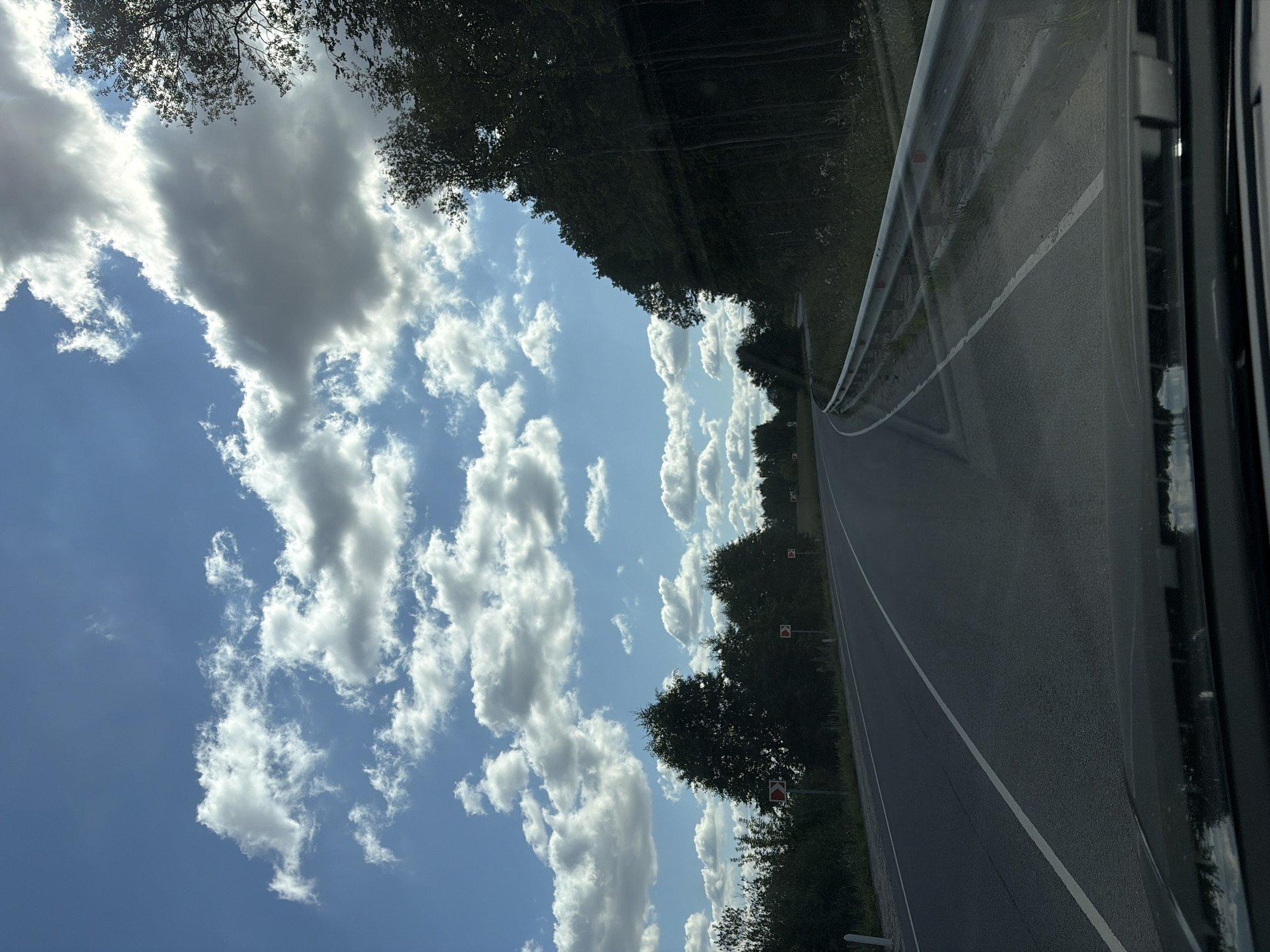The image depicts a road in the city of Kolontaevo, Russia, on a bright day with scattered clouds. The perspective is from inside a vehicle, looking out through the windshield. The road curves to the left, with a white line marking the lane. On the right side of the road, a metal guardrail runs along the edge, with vegetation growing around it. On the left side, there are trees and roadside signs indicating sharp turns. The sky is a vibrant blue with large, fluffy white clouds and some darker, shadowed clouds. Sunlight is breaking through the clouds in some areas. The overall impression is of a scenic drive through a rural or semi-rural area.