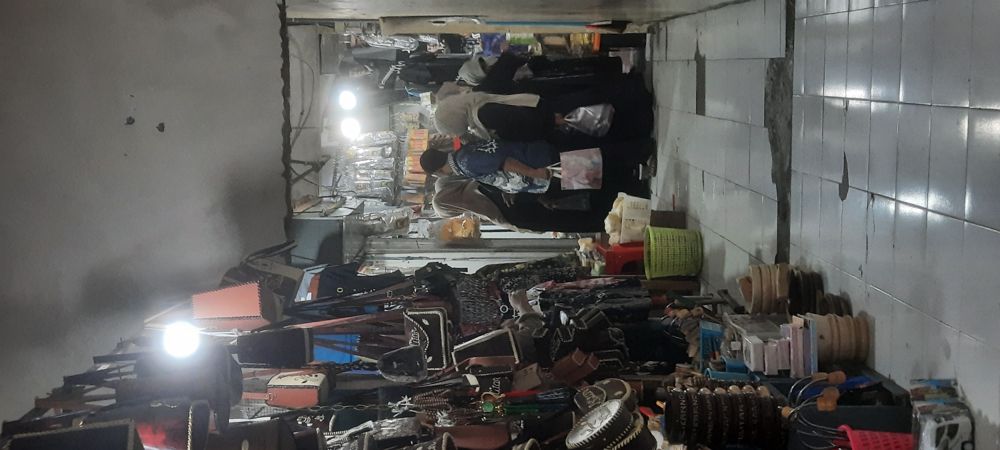 The image depicts an indoor market stall in Blitar, Indonesia, likely focused on selling bags and accessories. A variety of bags, made from different materials such as leather, are displayed hanging from the ceiling and on racks. Some bags are plain, while others have decorative elements or patterns. There are also smaller accessories like tassels and beaded items visible.
In the background, a group of people are gathered near a stall. Several individuals are wearing headscarves, suggesting a conservative cultural setting. One person, a male, is wearing a blue patterned shirt and appears to be interacting with the other shoppers. Two women, also wearing headscarves, are holding shopping bags. The stall behind them is filled with various items, possibly electronics or small personal accessories, displayed in clear packaging.
The environment appears to be a narrow passageway within the market, with a tiled floor that shows some signs of wear and tear. Lighting in the scene comes from overhead bulbs, casting a warm glow on the merchandise and the shoppers. To the left of the frame, more merchandise is stacked, including wooden items that appear to be hand-carved and some boxed items. A red basket is also visible. The overall impression is of a bustling, somewhat crowded market setting.