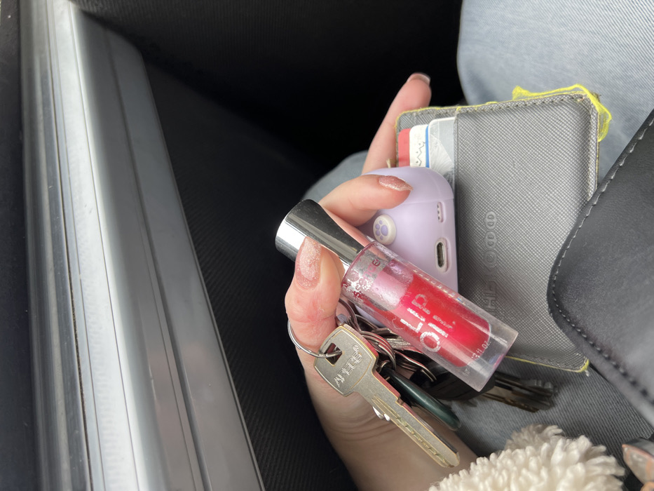 This image depicts a close-up view of a person's hand inside a vehicle in the city of Липицы, Russia. The hand, exhibiting light-colored, possibly glittery, nail polish, is holding a keyring with several keys, including a prominent brass-colored key labeled "APEX." Directly below the fingers, a clear cylindrical bottle containing red liquid, labeled "essence LIP OIL," is held upright. The hand and the held objects rest upon a dark gray textured wallet or cardholder, from which a bright yellow string or tag is partially visible, along with multiple cards inserted into its slots. Partially obscured behind the lip oil bottle and resting on the wallet is a small, light purple electronic device with a paw print design on its top surface and a USB-C port at its base. The person is wearing light blue jeans, visible in the lower right of the frame, and a fluffy white material is present in the lower left. The background shows the interior of a vehicle, specifically a car door panel with a light grey trim and a dark textured surface.