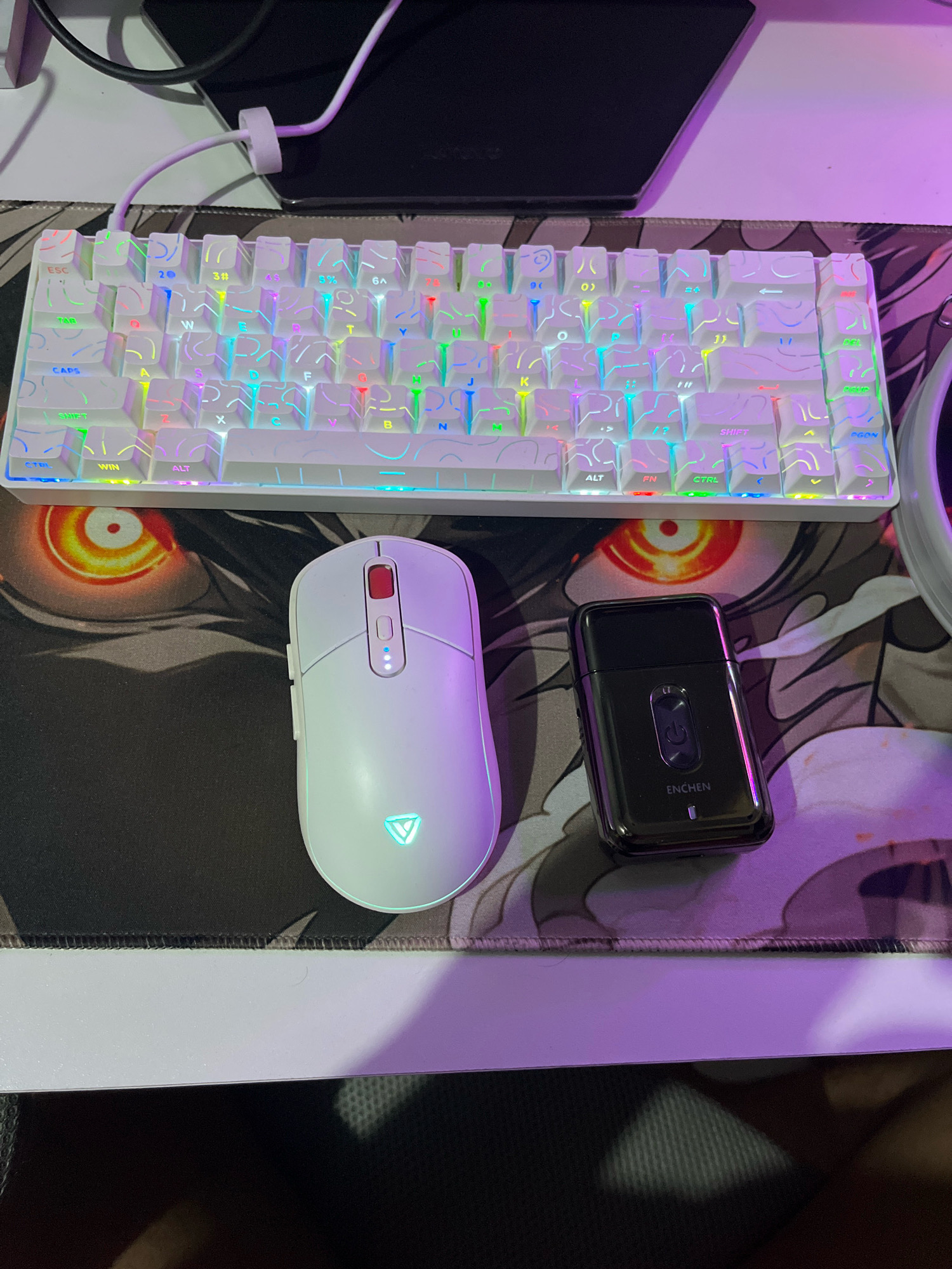 A white mechanical keyboard with RGB lighting sits on a black mousepad with an anime-style design. To the right of the keyboard is a white wireless mouse with a red scroll wheel and a glowing green logo. To the right of the mouse is a small black electric shaver with the brand name "Enchen" on it. The scene appears to be a desk setup, possibly in a home or office environment. The lighting is predominantly from the RGB keyboard, casting colorful hues onto the surrounding objects and the mousepad. There is no indication of the location being in Cimahi, Indonesia.