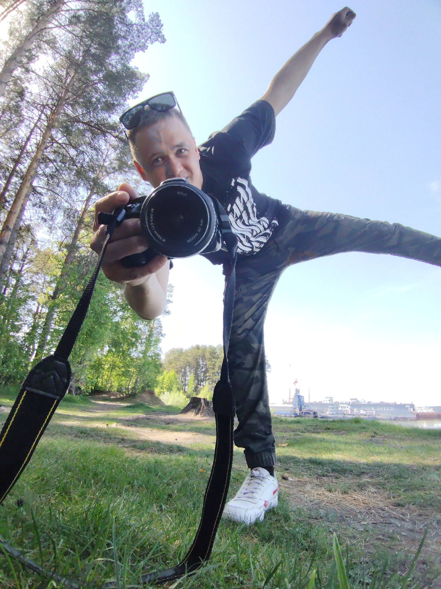 A man poses with a Nikon camera in an outdoor setting, likely in Chaikovsky, Russia. He is positioned on grass, with one leg extended to the side and one arm raised in the air. He is wearing sunglasses on his head, a dark T-shirt, camouflage pants, and white sneakers. The camera strap is visible around his neck. In the background, there is a body of water, a ship, and industrial structures. Trees surround the scene.