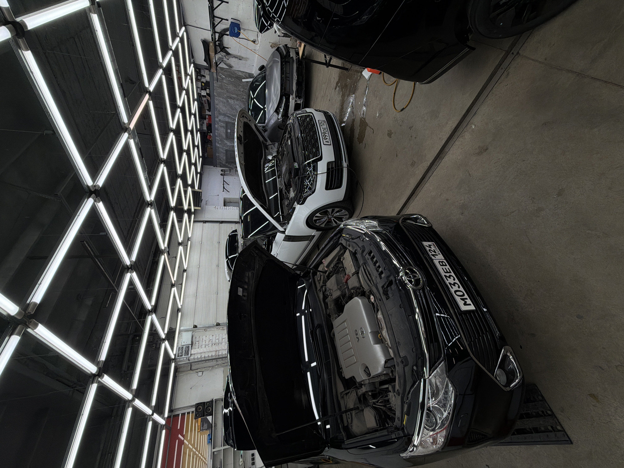 The image shows an interior space, likely a car detailing or repair shop. The prominent feature is an array of bright, grid-like LED lighting fixtures on the ceiling. Several cars with their hoods open are arranged in the space. One car in the foreground is black with a license plate that reads "MO33EB 124 RUS", and the second car is white with a license plate "A999OE 124". The location is indicated by the license plates as Krasnoyarsk, Russia. The floor appears to be concrete. No people are visible in the frame.