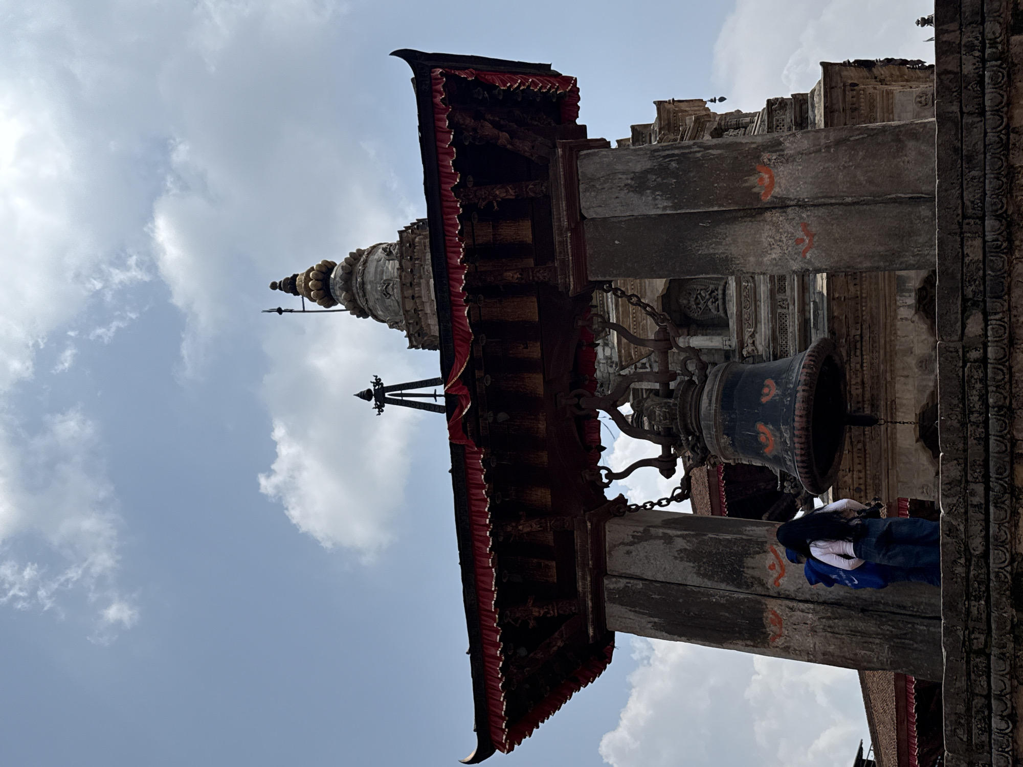 The image shows a view of a temple structure in Bhaktapur, Nepal, under a partly cloudy sky. The focus is on a multi-tiered pagoda-style roof with red fabric trim, supported by weathered stone pillars. A large bell, suspended by chains, hangs between the pillars. The stone pillars feature carved details and orange markings. Part of a person wearing a blue shirt, a white shawl and blue pants can be seen at the bottom-right corner of the image. The sky is pale blue with white clouds.