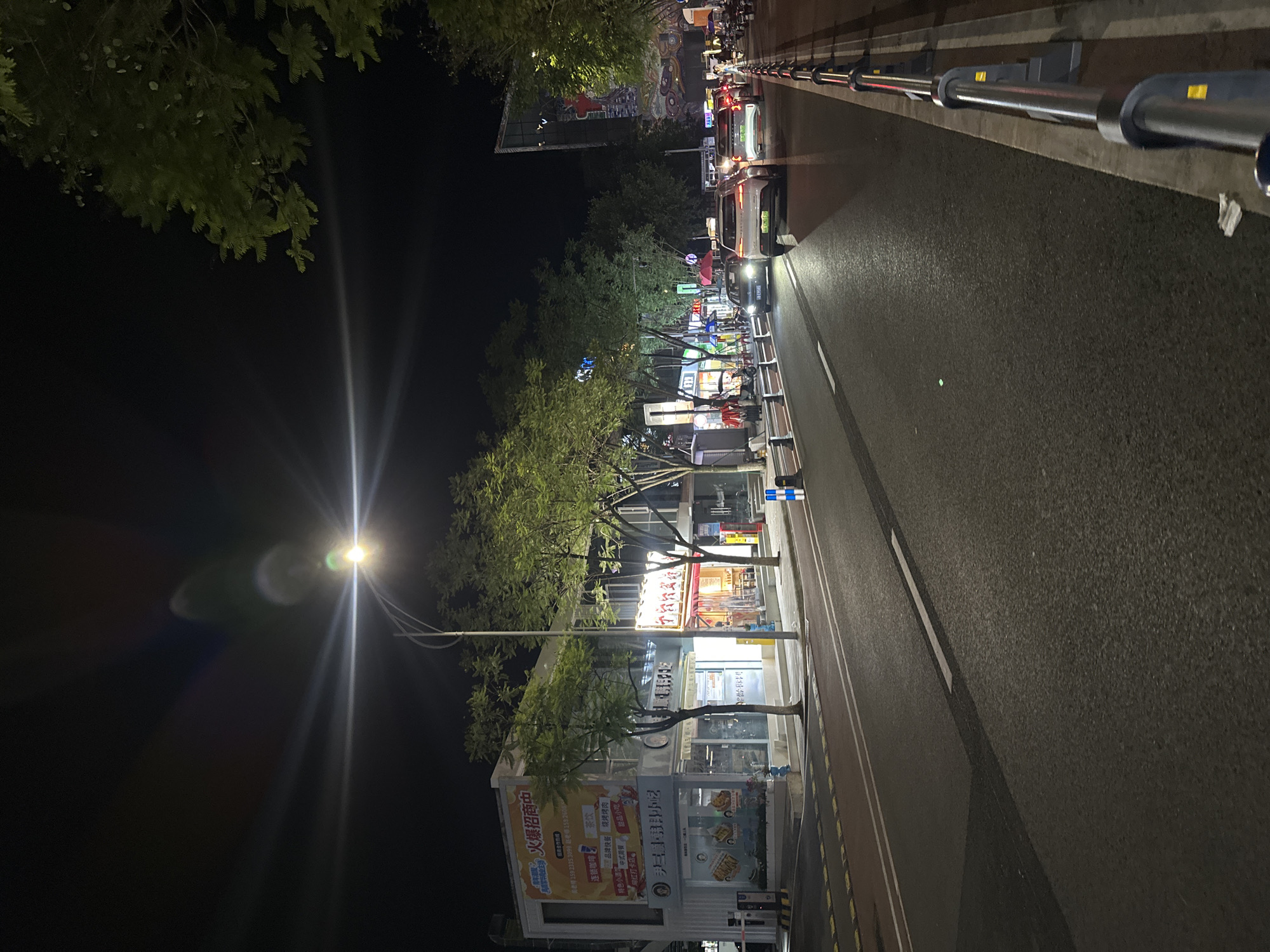 This is a night scene on a city street, likely in the Chongqing High-tech Zone, China, as indicated by the presence of various businesses and traffic. The street is paved with asphalt and marked with white lines, indicating lanes and pedestrian crossings. A metal railing with black and grey supports runs along the right side of the street, separating the road from the sidewalk. Several vehicles, including cars and possibly motorcycles, are visible on the road, some driving and others parked.
On the left side of the street, there are several buildings with illuminated storefronts. One building has a prominent banner with Chinese characters that appears to be an advertisement for business opportunities. Other storefronts show signs of commercial activity, with interior lighting and displays visible through the windows. Trees with green foliage line the sidewalk, providing some greenery amidst the urban environment.
A street lamp illuminates the scene, casting a bright light that creates lens flares in the camera. The sky is dark, indicating that it is nighttime. The overall impression is of a bustling commercial area at night, with ongoing economic activity. The presence of modern vehicles and buildings suggests a developed urban area.