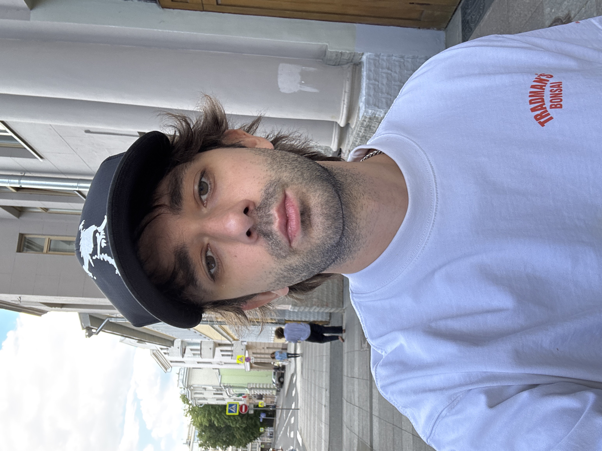 The image is a self-portrait of a man wearing a black baseball cap and a white t-shirt with "TRADMAN'S BONSAI" printed in red. He has dark hair and stubble. He is standing outdoors on a sidewalk in a city. Buildings line the street, and there are traffic signs and a crosswalk visible. The sky is cloudy, suggesting it is daytime. The overall impression is casual and candid.