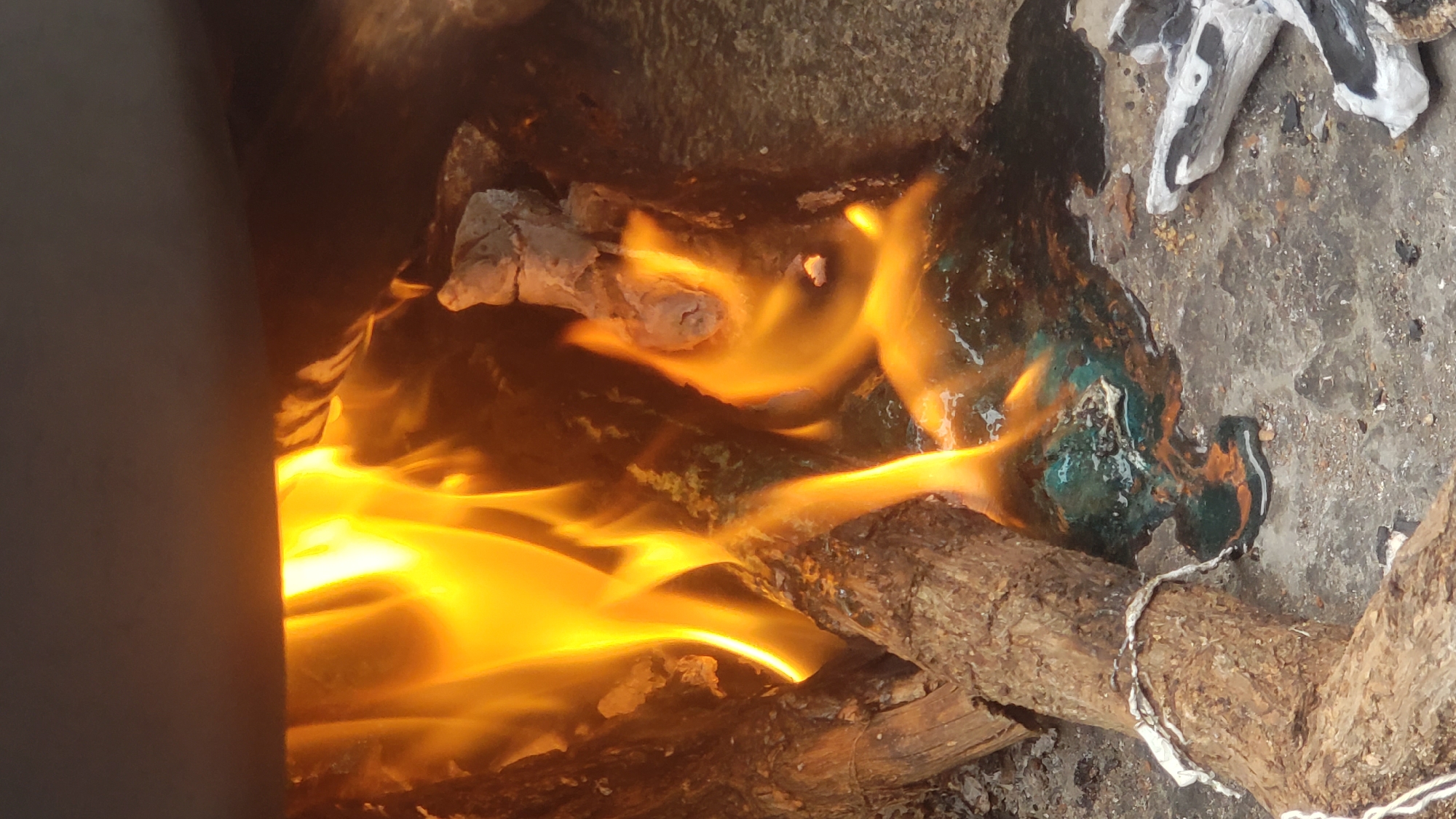 The image shows a close-up view of a fire burning within what appears to be an outdoor setting, possibly a fire pit or an improvised stove in Sabon Tasha, Nigeria. The flames are a vibrant orange and yellow, licking at pieces of wood and what seems to be a dark, viscous liquid that has melted or spilled onto the rough, grey ground. There are also some white and grey charred remnants of wood or other materials scattered around the burning area. The scene is dimly lit, with the fire being the primary source of illumination, casting a warm glow on the surroundings. The overall impression is one of a simple, perhaps rural, fire being used for some practical purpose, with the materials burning suggesting a makeshift setup.