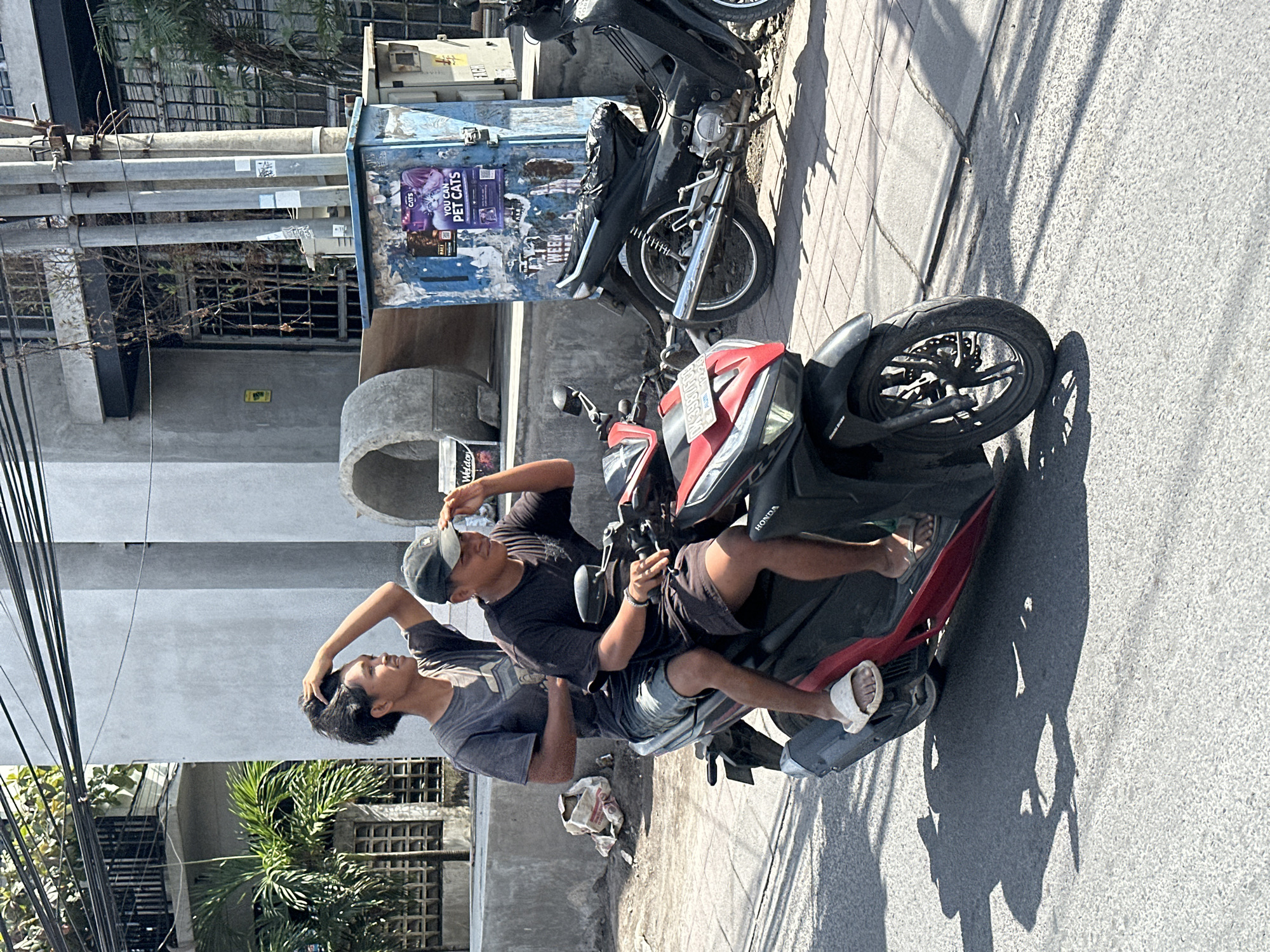 Two young men are riding a red and black scooter on a paved road in North Kuta, Indonesia. The man in the front, wearing a black t-shirt and black shorts, is driving the scooter and shielding his eyes with his hand, appearing to be looking at something in the distance. He is wearing a dark baseball cap. The man in the back, wearing a grey t-shirt and denim shorts, has his hand on his hair. Both men are wearing sandals. The scooter is a Honda. In the background, there is a light grey building with a barred window and palm trees. A blue electrical box with posters on it is visible on the right side of the image. Other parked motorcycles are also present. The lighting suggests it is daytime.