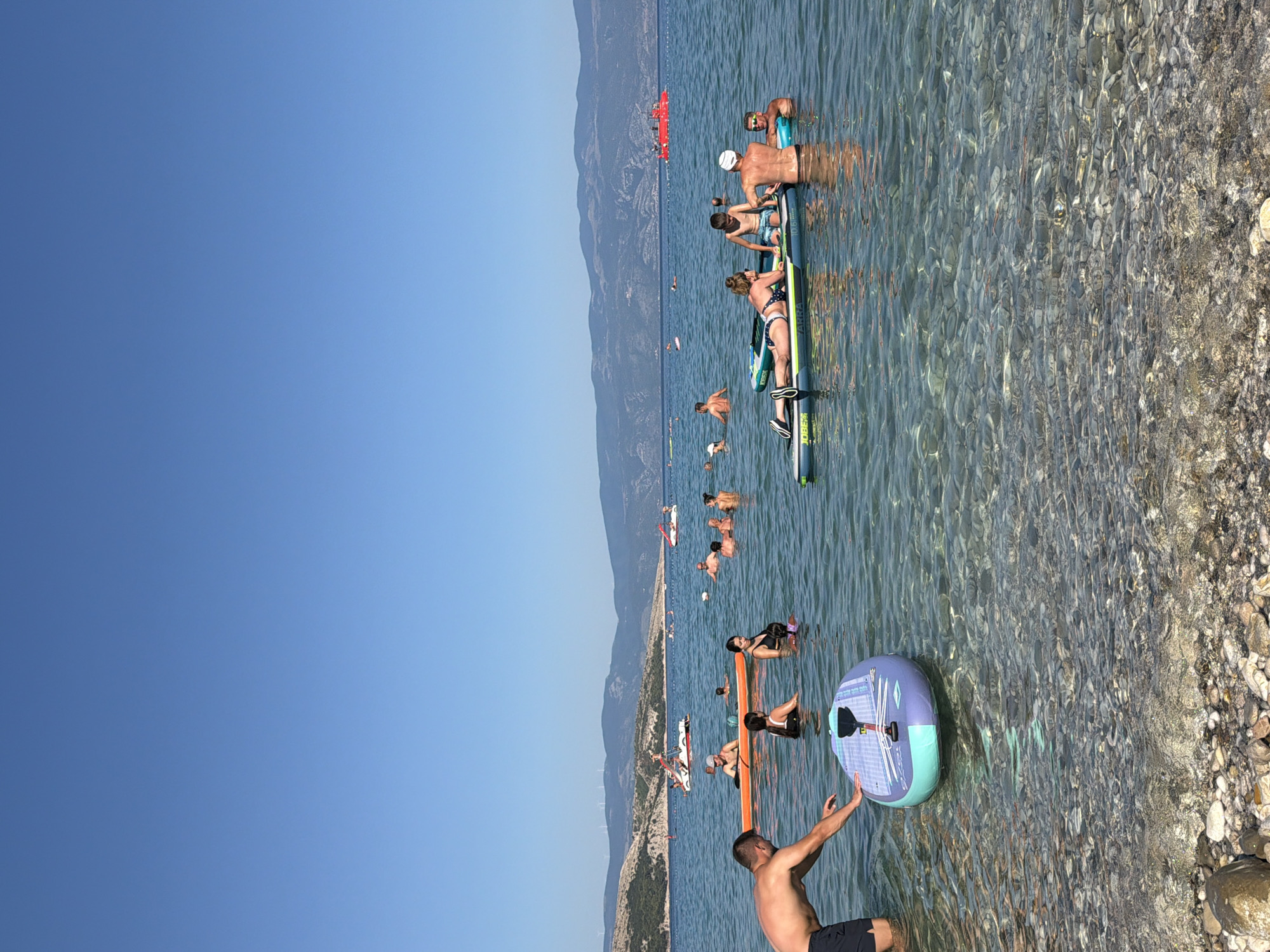 The image depicts a crowded beach scene in Općina Baška, Croatia, on a clear, sunny day. The water is calm and clear, revealing a rocky seabed. Several people are engaged in various water activities, including swimming, sunbathing on inflatable boards, and paddling on paddleboards. In the foreground, a man in black shorts is partially submerged in the water, with a paddleboard in front of him. To the right, a group of people are on two paddleboards, with a young boy sitting between two adults. Further out in the water, other individuals and groups are visible, some on paddleboards and others simply swimming. A bright red boat is present in the distance, along with a white motorized watercraft. The background features a hilly coastline with scattered vegetation and mountains. The sky is a clear, deep blue.