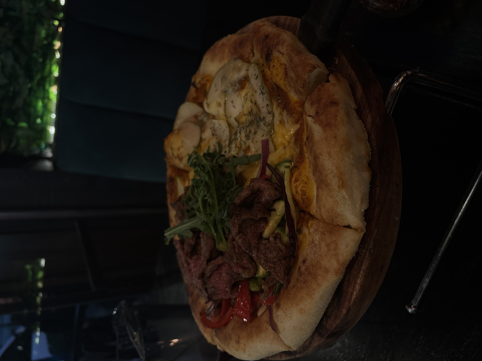 A tilted eye-level shot shows a wood-fired pizza on a wooden platter, likely in a restaurant, possibly in Mexico City, Mexico. One half of the pizza is topped with chunks of meat, arugula, red peppers, and green peppers; the other half has creamy cheese and herbs. A black server spoon rests against the top of the pizza. To the left, a blurred scene includes artificial plants and dark furniture. To the right, a dark environment with a small, metallic object.