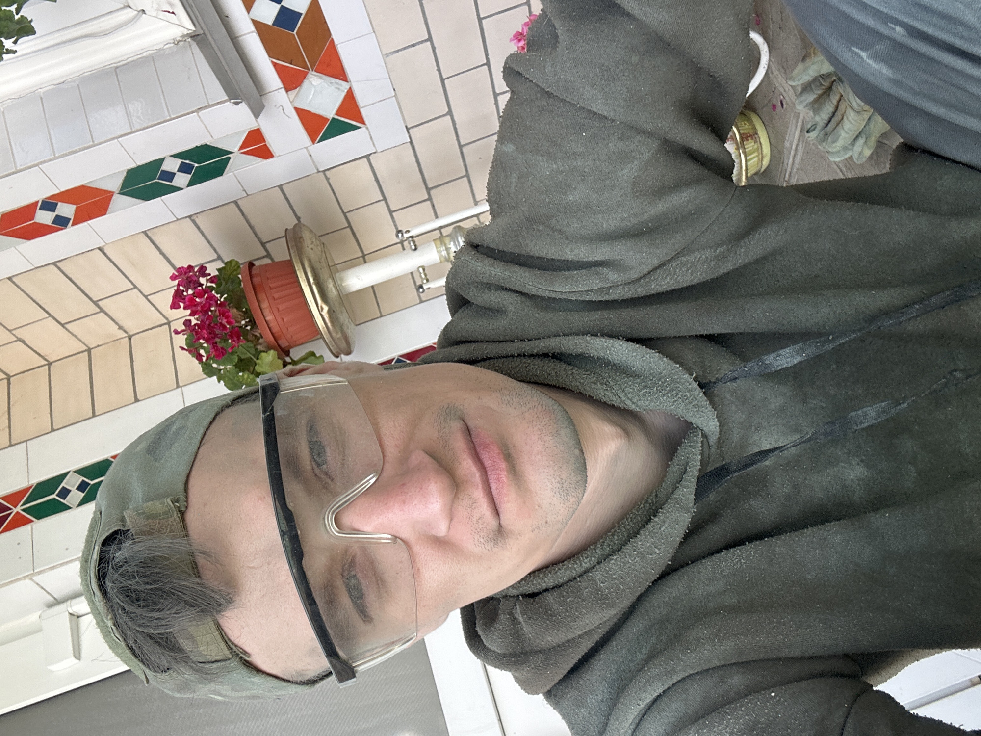 A man wearing a green baseball cap and safety glasses is taking a selfie. He has on a green hooded sweatshirt. The background shows a tiled wall with colorful geometric patterns, a potted plant with pink flowers on a stand, and some white trim. The overall scene suggests an outdoor or semi-outdoor location, possibly a porch or balcony.