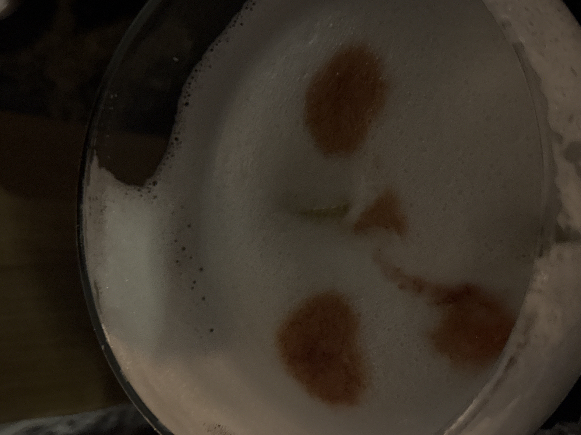 An overhead, close-up shot captures the surface of a drink in a glass, set against a dark, out-of-focus background. The beverage is topped with a thick layer of dense, white foam composed of numerous small bubbles. Irregularly shaped spots of dark reddish-brown liquid are artfully dotted across the foam, creating a decorative pattern. A faint hint of a small, bright green element is visible beneath the foam's surface, possibly indicating a garnish. The rim of the clear glass is partially visible along the left and right edges of the frame. The lighting is dim, suggesting an evening or nighttime setting indoors. No people, discernible activities, or visible text are present in the image, which focuses purely on the visual texture and composition of the drink. The scene is located in Ramat Gan, Israel.