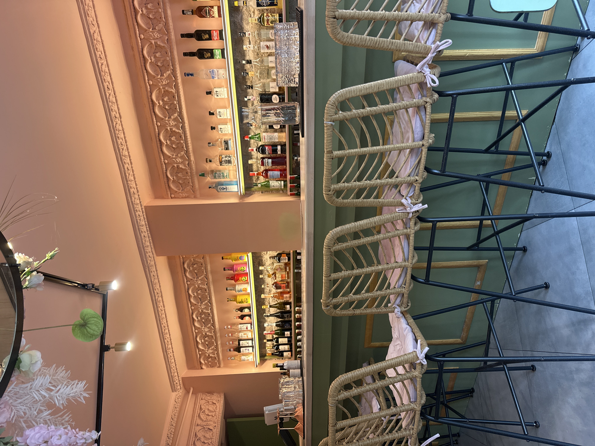 The image depicts the interior of a bar or restaurant, with a focus on the bar area and seating. Behind the bar, shelves are stocked with a variety of liquor bottles, illuminated by warm lighting. On the bar counter, glassware and a carafe of water are visible. In the foreground, several woven bar stools with light pink cushions are arranged facing the bar. The walls are painted a soft pink above and a muted green below, with ornate gold-colored frames on the lower portion of the wall. Decorative floral elements hang from the ceiling. There are no people visible in the image. The scene is clean and well-lit, suggesting an operational establishment. The image does not provide any specific visual cues that would indicate its location in Ternopil, Ukraine.