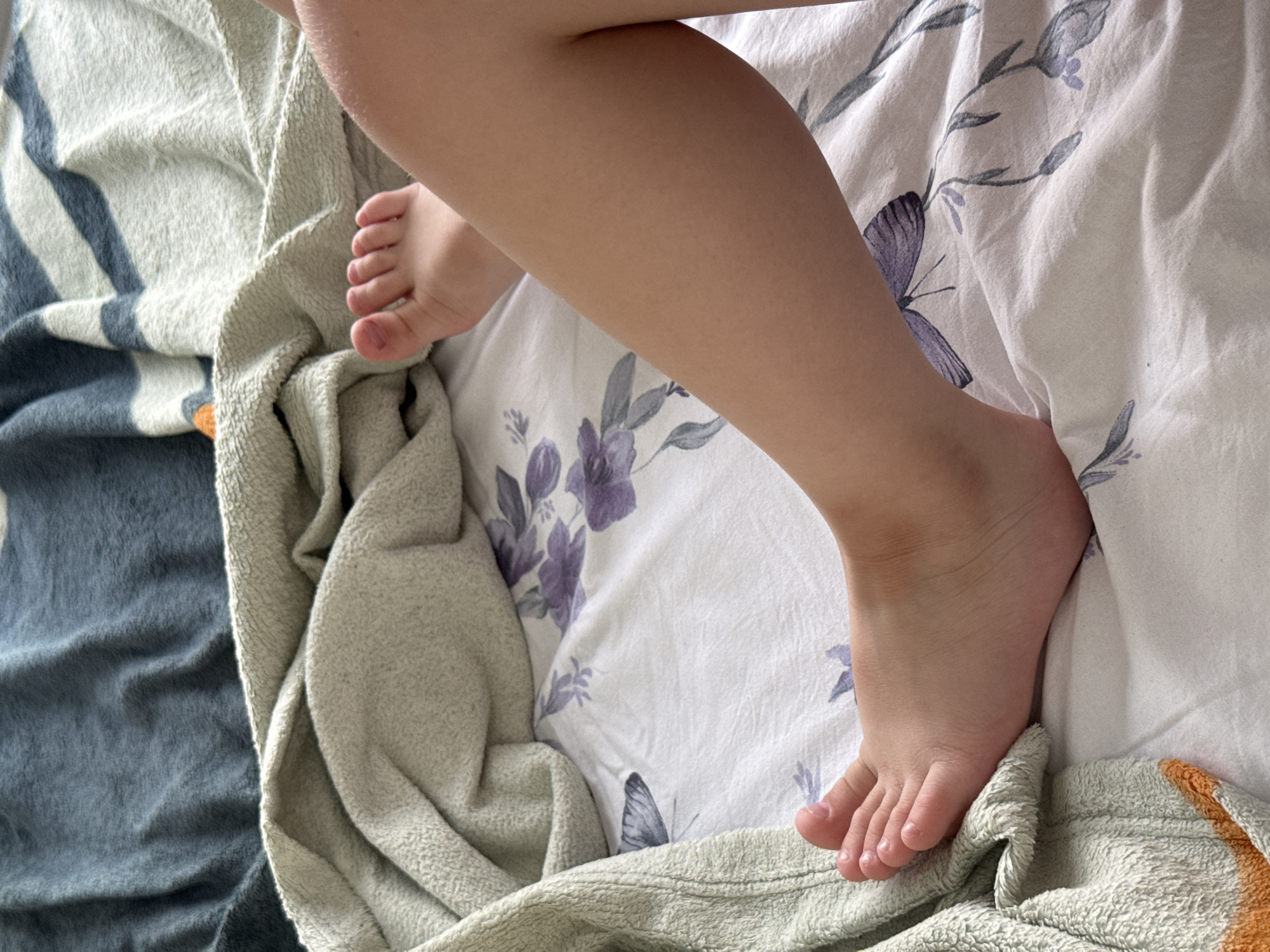 The image shows a close-up of a person's legs and feet resting on a bed. The bedding has a floral pattern with purple flowers and butterflies. A textured blanket in shades of blue and green is draped over the bed. The scene is indoors, with natural light suggesting a daytime setting. There is no indication of the location being in Lyubertsy, Russia.