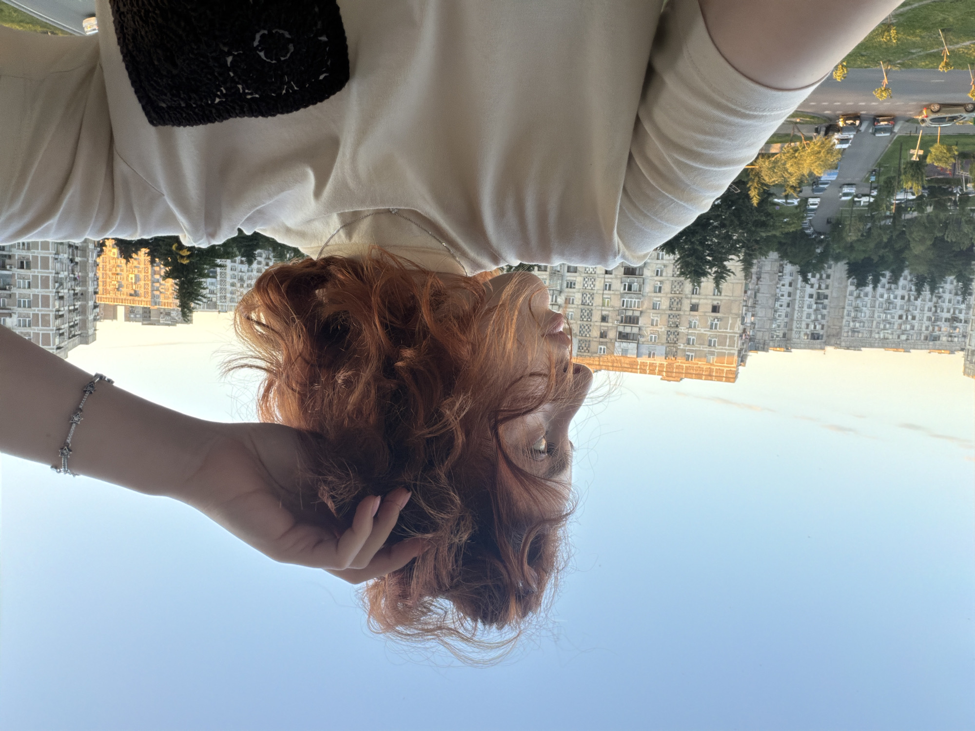 A woman with red curly hair is holding her hair with her left hand in an upward-looking selfie. She is wearing a light-colored shirt or sweater with a dark brown square pattern on the upper left. The woman is also wearing a silver bracelet on her left wrist. In the background, there is a clear blue sky, multi-story buildings, trees, parked cars, and a road. The buildings suggest an urban area in Tbilisi, Georgia.