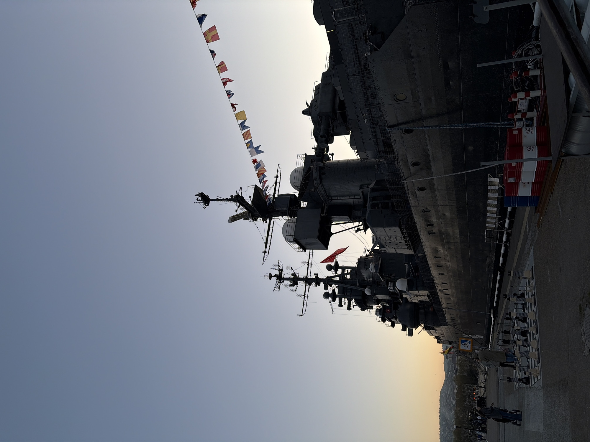 A warship is docked in Novorossiysk, Russia. The ship is gray and has a variety of antennas and radar equipment on its masts. A line of colorful flags is strung above the ship. There are a few people milling around on the dock next to the ship. The sky is pale and clear. A cityscape with trees is in the background on the right of the frame. There are some barrels next to the ship.