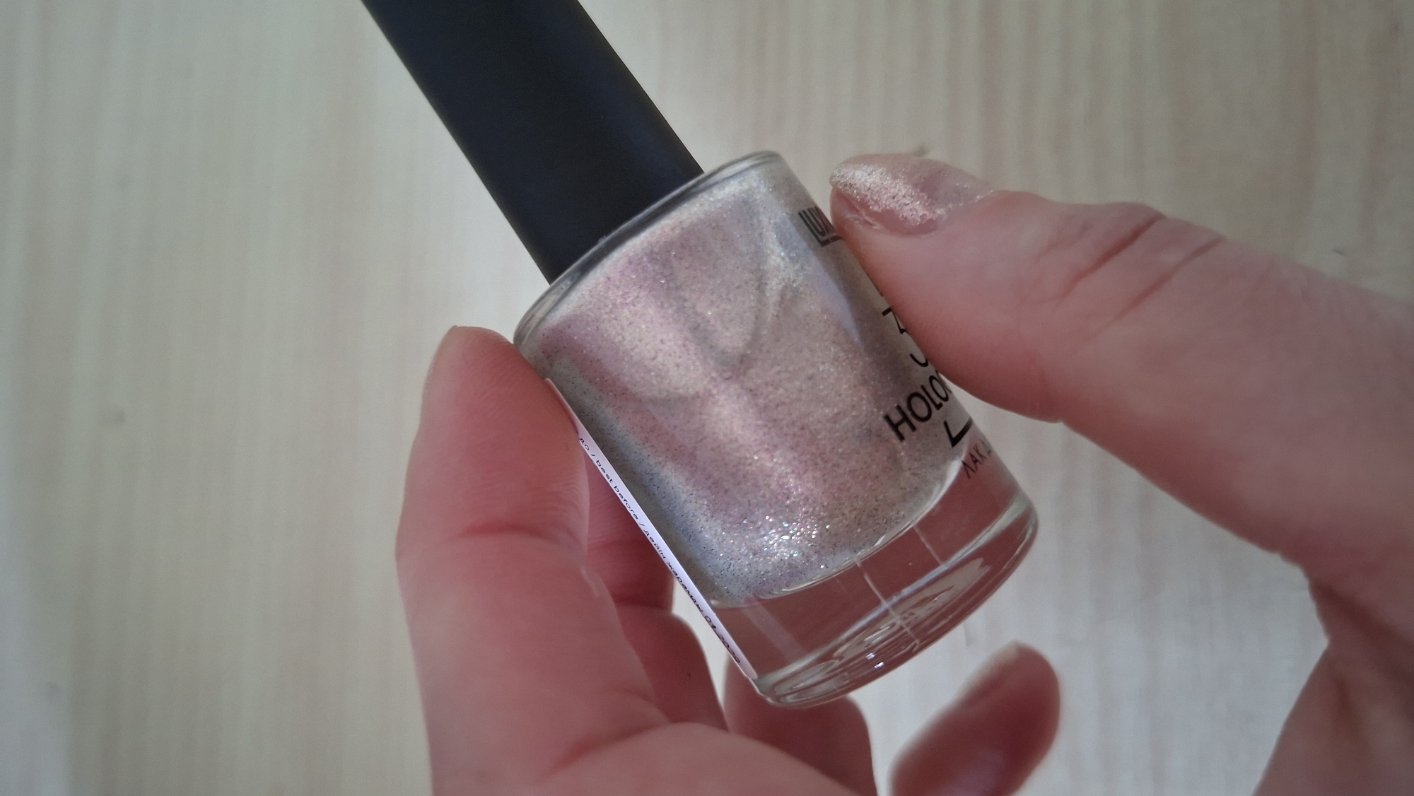 A hand holds a bottle of holographic nail polish against a light-colored wood surface. The bottle is mostly clear with a black cap. The nail polish appears to be a pale, shimmering color. The hand also has nail polish applied. The location is unknown.