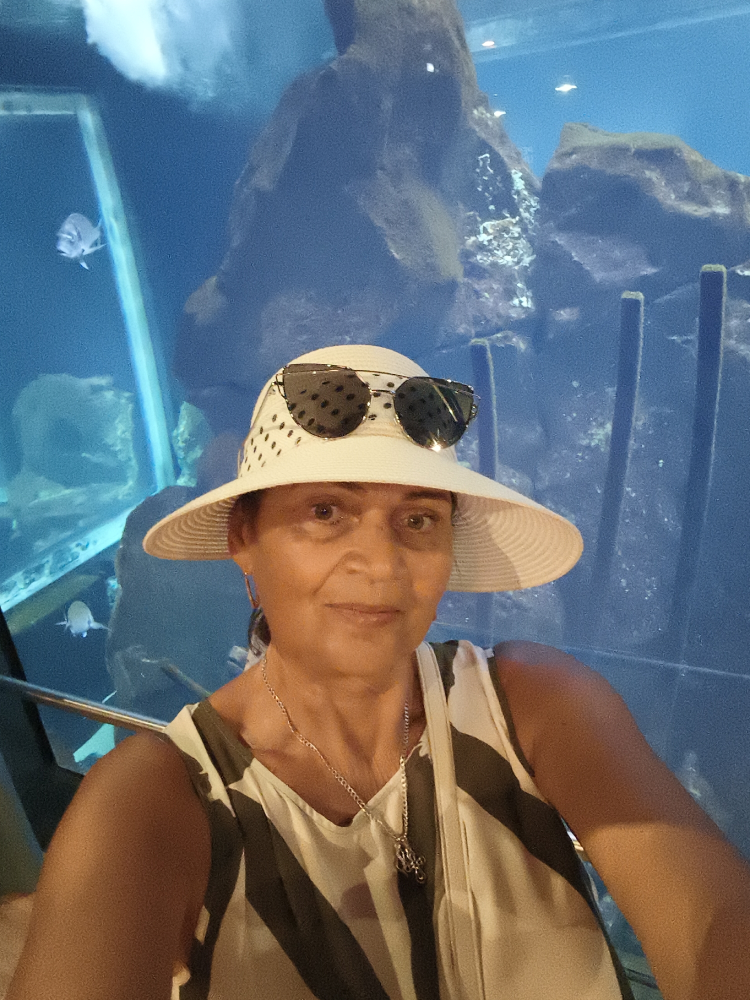 A middle-aged woman with brown skin and dark hair wears a white, wide-brimmed hat with black polka dots and a pair of dark sunglasses. She is looking at the camera and smiling slightly. She is wearing a sleeveless top with a pattern of green and white stripes, and a silver necklace with a pendant. She is standing in front of a large aquarium with blue water and large rocks. There are a few fish visible in the aquarium. The lighting is dim. The location is the Livorno Aquarium in Livorno, Italy.