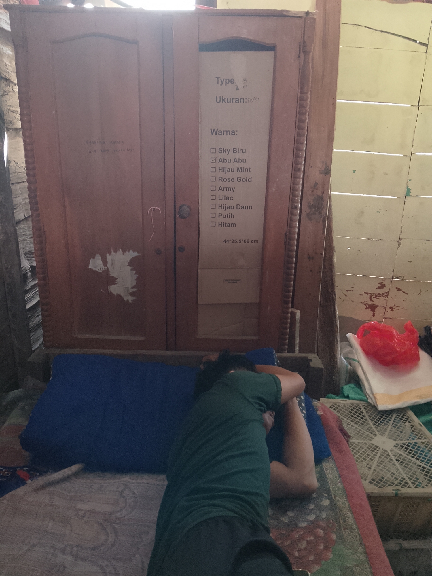 The image shows a room with a wooden cabinet. On the cabinet door, there is a handwritten label with text in Indonesian. Below the cabinet, a person is lying down on a blue pillow and a patterned mat. The room appears to be a residential interior in Kasiman, Indonesia, characterized by its rustic wooden furniture and wall constructions. There are also some plastic bags and a fan visible in the background.