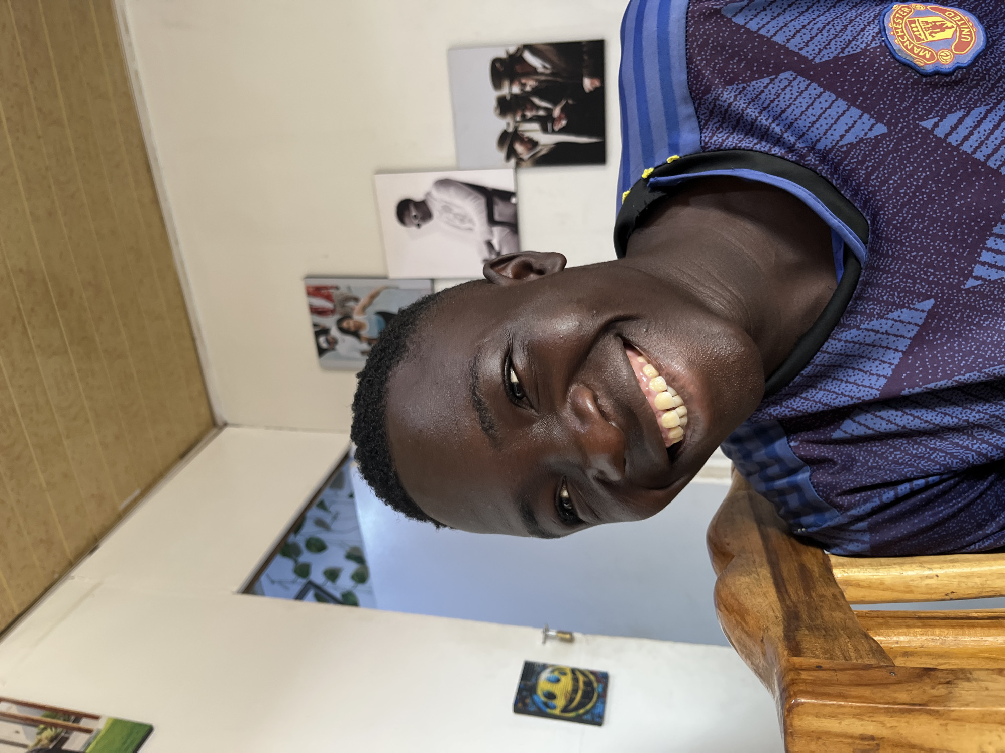 The image shows a young man wearing a Manchester United soccer jersey, sitting on a wooden chair. He is smiling broadly. Behind him, on the wall, are several framed photographs. The setting appears to be indoors, possibly in a residential area. It is impossible to determine if the location is in the city of Jos, Nigeria from this image.