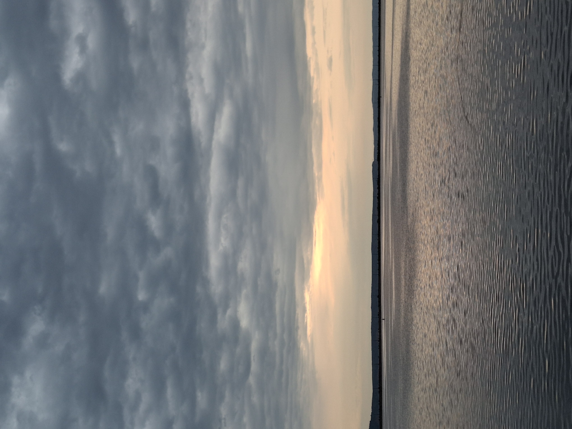 The image depicts a body of water next to a land strip with a dramatic sky above. The water has a textured surface, suggesting ripples or small waves. The land strip is dark and appears to have some minor vegetation. The sky is mostly cloudy with a transition to a lighter, possibly sunset-lit area closer to the water. The photograph is taken horizontally. The location is Chaikovsky, Russia.