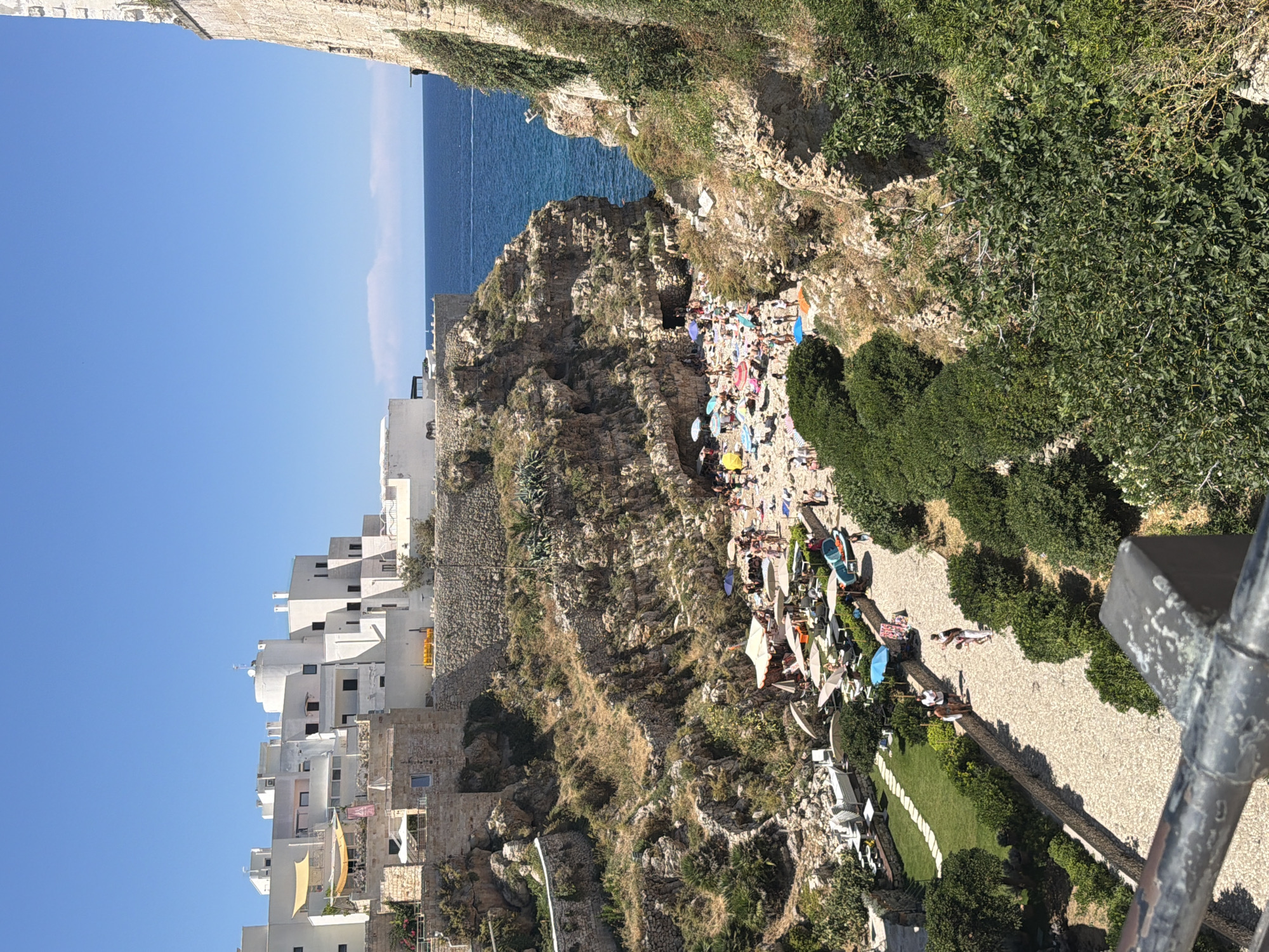 This is a long shot, high-angle, medium-light, outdoor, daytime photograph of a beach cove in Polignano a Mare, Italy. The scene is dominated by a rocky cove with a beach packed with people. White buildings are visible on the cliffs above the cove, as well as a stone wall and abundant greenery. The water is a deep blue.