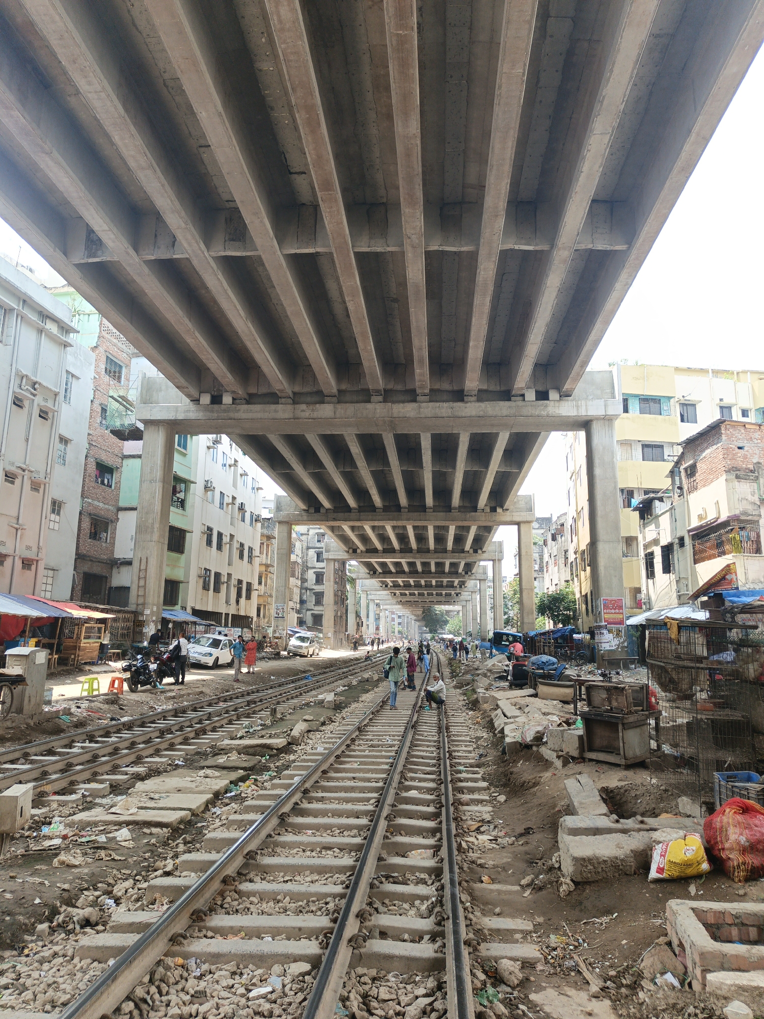 The image shows a railway track running through a congested urban environment in Dhaka, Bangladesh. Above the tracks, a large concrete overpass structure dominates the scene. Buildings of varying heights and conditions flank both sides of the tracks, with a mix of residential and commercial establishments. Along the sides of the railway, there are makeshift stalls and vendors. People are walking on and alongside the tracks. Litter and debris are scattered throughout the scene. The overall impression is one of urban density and activity.
