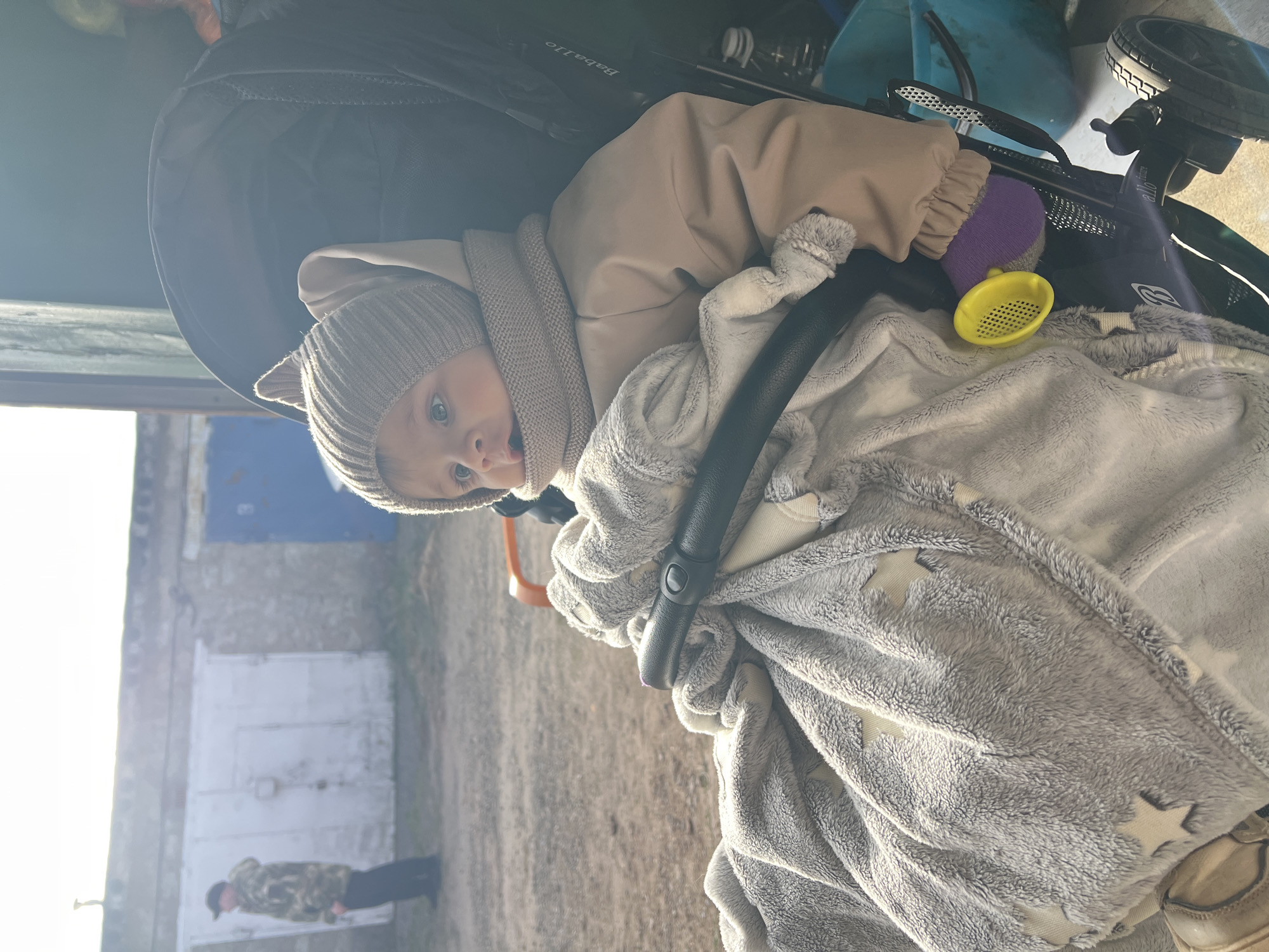 A baby is lying in a stroller, wrapped in a light gray blanket with white stars. The baby wears a beige jacket and a gray knit hat. The stroller has a black frame, a yellow and purple toy, and a beige neck protector. The background shows a building with a blue door and a person standing in the distance. The image was taken in Noviy, Russia.