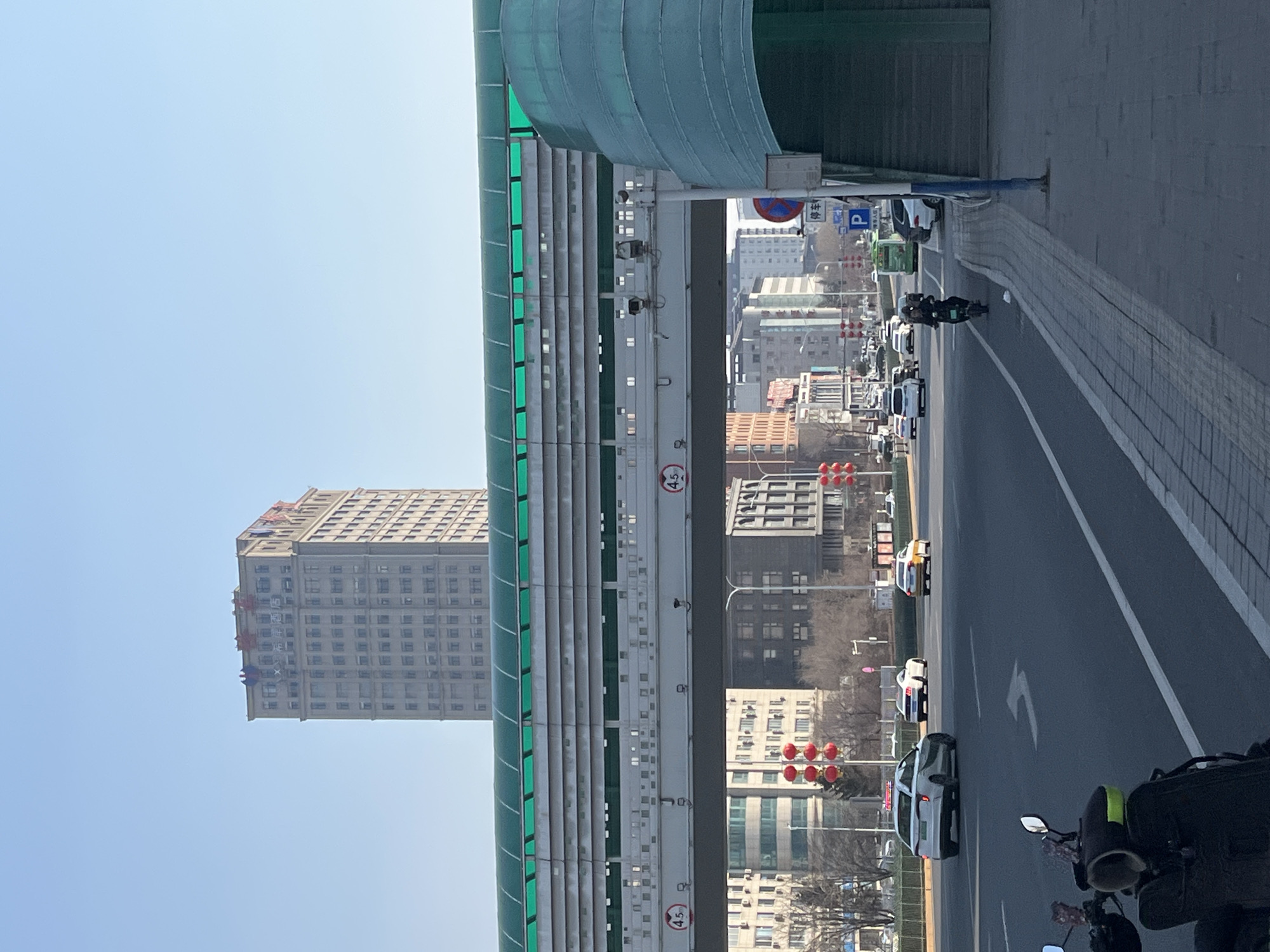 This image depicts a street scene in Changchun City, China, specifically at the intersection of Renmin Street and Xinfa Road, with the Xingfu Hotel visible in the background. The scene is observed on a clear, bright day.
In the background, a tall, multi-story, light grey building, identified as the Xingfu Hotel by red Chinese characters (幸福饭店) on its upper facade, dominates the skyline. In the mid-ground, a pedestrian overpass with a green, transparent curved roof and grey paneled sides spans horizontally across a multi-lane asphalt road. The overpass structure includes two circular signs indicating a 4.5-meter height clearance (4.5米) for vehicles below. On the right side, an enclosed, curved staircase with matching green transparent panels provides access to the overpass.
The multi-lane road features white lane markings, including a white left-turn arrow painted on the pavement in the foreground. Several vehicles are in motion: a white sedan is turning left, a white sports car and a yellow taxi are further down the road, and a person rides a dark scooter/moped in the rightmost lane. A green bus is visible in the distant traffic.
On the right, a tiled sidewalk runs parallel to the road. Adjacent to the overpass staircase, a blue rectangular sign with a white 'P' for parking and the Chinese characters "停车" (parking) is mounted, alongside a red circular no-parking sign. On the left side of the road, traffic lights with multiple red spherical lanterns are visible. Decorative red lanterns are also suspended from poles along the street, suggesting a festive ambiance. Bare trees line the street, indicating a season without foliage. In the bottom left, a dark-colored scooter or moped is partially visible, likely stationary.