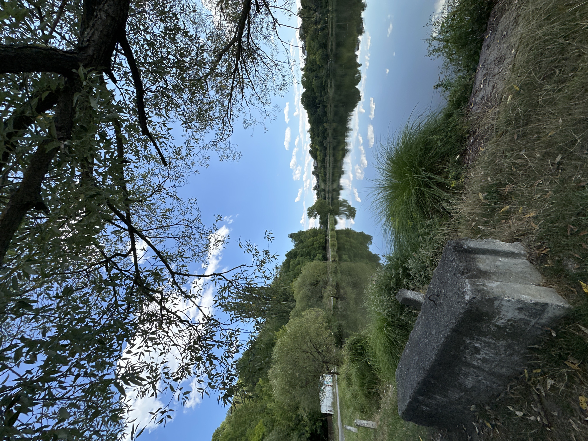 This image depicts a serene natural scene by a body of water, likely a lake or river, in Makiv, Ukraine. The foreground is dominated by a large, weathered concrete block placed on a grassy embankment. Tall, lush green grass surrounds the block, with dry leaves scattered on the ground. The water's surface is calm and reflective, mirroring the bright blue sky dotted with fluffy white clouds and the dense green foliage of trees on the opposite bank.
The trees form a verdant backdrop, creating a natural landscape. Some of the trees have branches extending into the frame from the top, with dappled sunlight filtering through their leaves. The overall atmosphere is peaceful and tranquil, capturing a moment of natural beauty in a presumably rural or semi-rural setting within Makiv. There are no people or animals visible in the image. The concrete block suggests human presence or past activity, perhaps related to infrastructure or a public space.