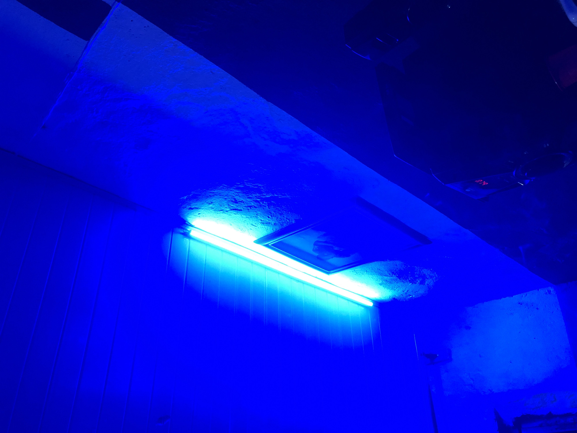 The image displays an indoor scene in the city of Unknown, Unknown, characterized by a dominant blue artificial light source. A long, tubular light fixture, possibly fluorescent, extends diagonally across the upper-middle portion, emitting bright, cool white light that appears cyan-blue against the overall blue environment. This fixture is mounted on a rough, textured wall, possibly concrete or stucco. Directly below the light, a dark-framed object, likely a picture or mirror, is mounted on the same wall. The upper left section of the image reveals a horizontal-paneled surface, indicative of a ceiling or an upper wall section. In the lower right quadrant, a darker, less distinct area contains equipment, including an electronic device exhibiting a small display of red alphanumeric characters. The intense blue illumination creates deep shadows and highlights the textures of the surfaces. No individuals are present in the scene.