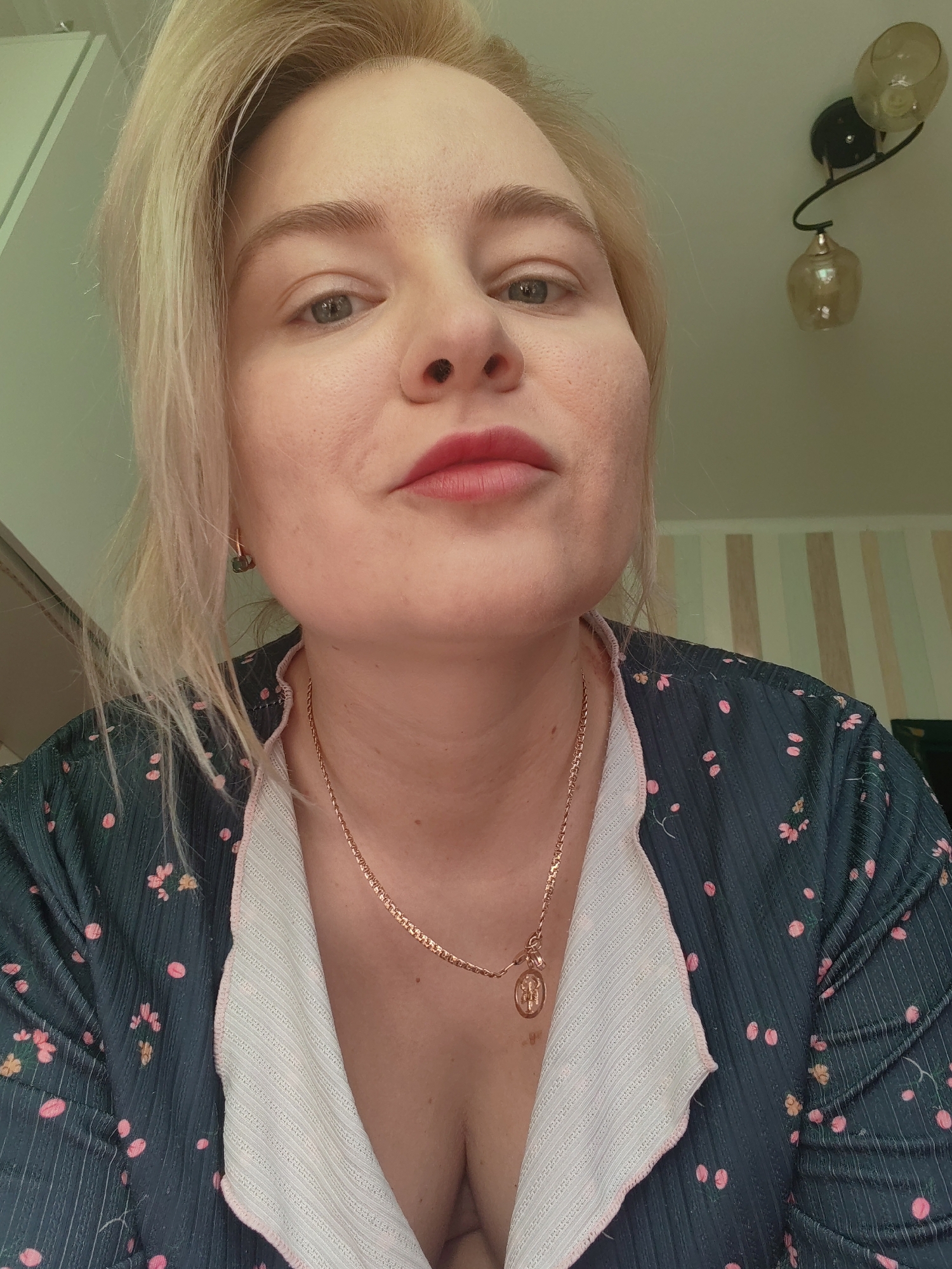 A fair-skinned woman with blonde hair is centered in the frame. She is wearing a dark blue floral top with a white, striped collar. A gold necklace with a pendant hangs around her neck. She is looking directly at the camera with a slight pursing of her lips. Behind her, a wall features vertical stripes and a decorative light fixture. There is a partial view of what appears to be a white cabinet in the upper left corner. The location in Izhevsk, Russia, cannot be determined from the information available in the image.