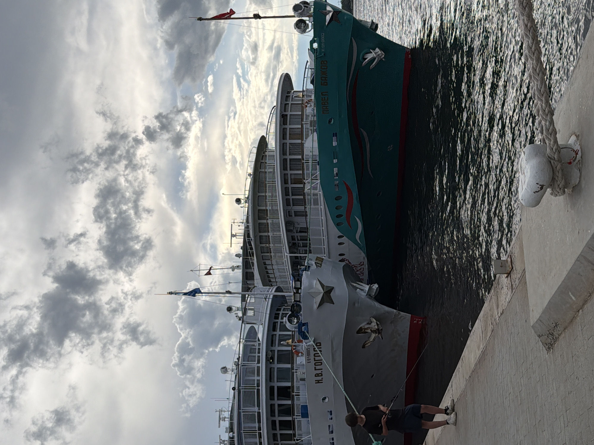 Two ships are docked side-by-side on a sunny day with scattered clouds. The ship in the foreground is gray and white with the text "Н.В. ГОГОЛЬ" visible on its side. It has a prominent star emblem on its bow and an anchor. Behind it, a teal-colored ship is docked, featuring the text "ПАВЕЛ БАЖОВ" and a decorative dragon-like motif. Both ships appear to be passenger vessels. A thick rope connects the gray ship to a white cleat on the concrete pier. On the pier, a person is visible in the lower right corner, appearing to be fishing with a rod. The water is calm with reflections of the ships. The sky is partly cloudy, suggesting it could be midday. The scene takes place at a dock, likely in Perm, Russia, given the ships' names and general aesthetic. The overall mood is tranquil and everyday.